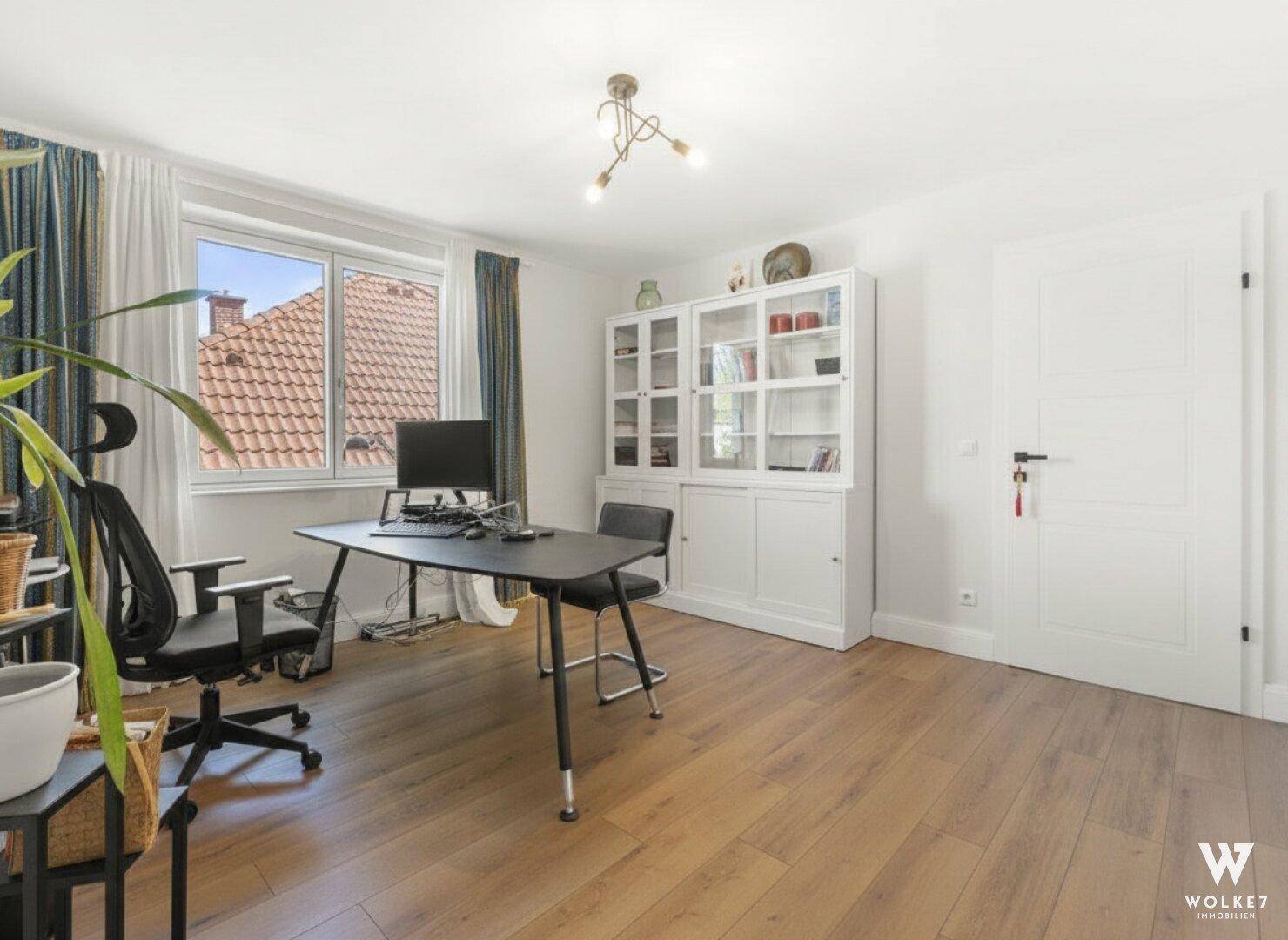 Helles Arbeitszimmer mit Holzboden, großem Fenster und Blick auf die Dächer.