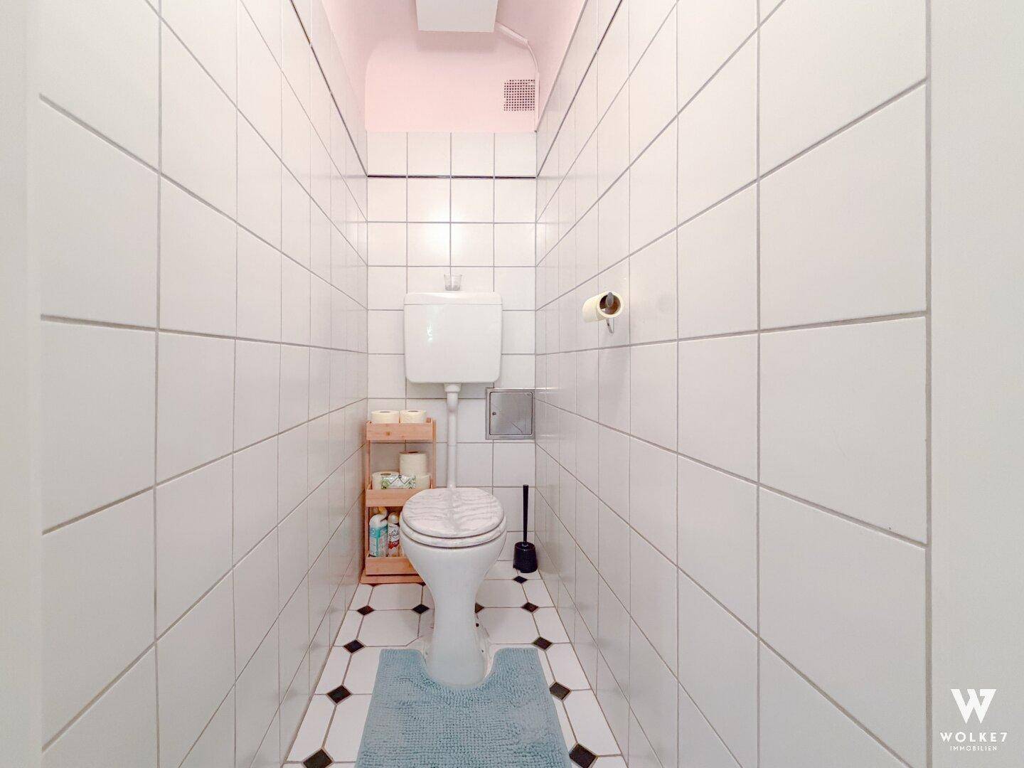 Separates WC mit weißen Fliesen, Steh-WC und kleinem Regal.