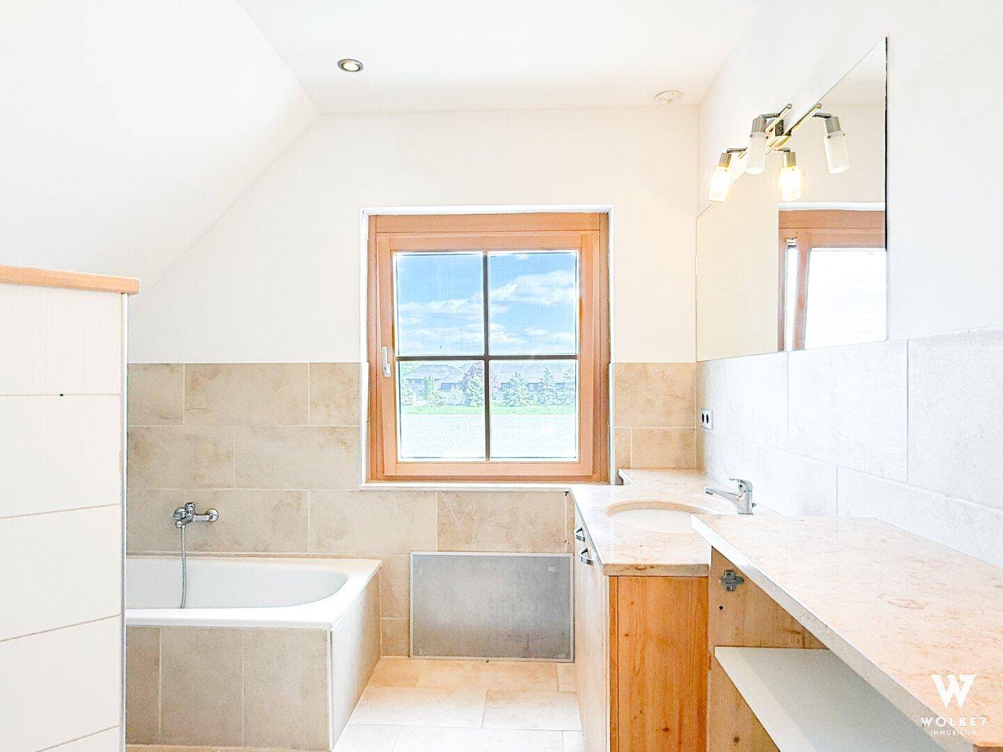 Helles Badezimmer mit Badewanne, Fenster und Holzwaschtisch.