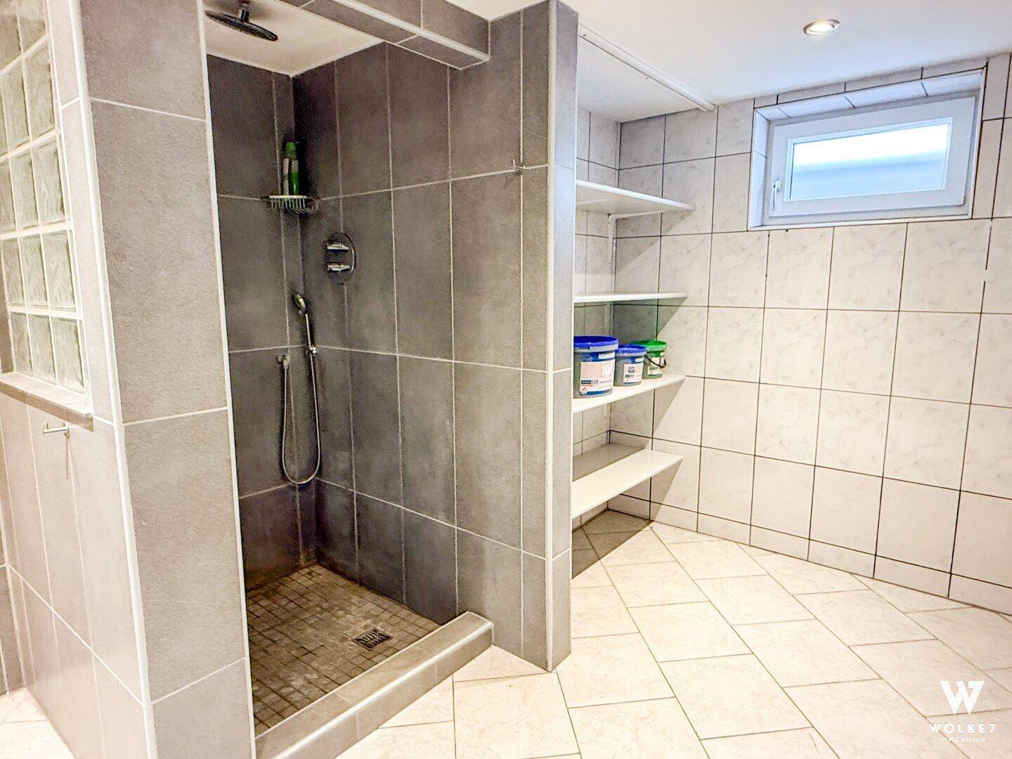 Graues Badezimmer mit gefliester Dusche, praktischen Ablagen und kleinem Fenster.