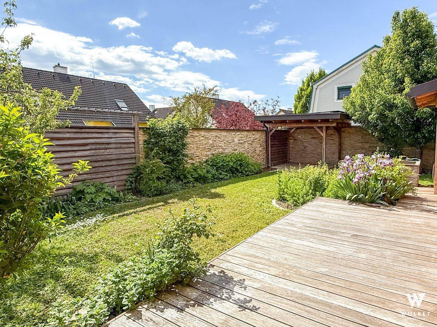 Sonniger Gartenbereich mit Holzterrasse, Rasenfläche und üppiger Bepflanzung.