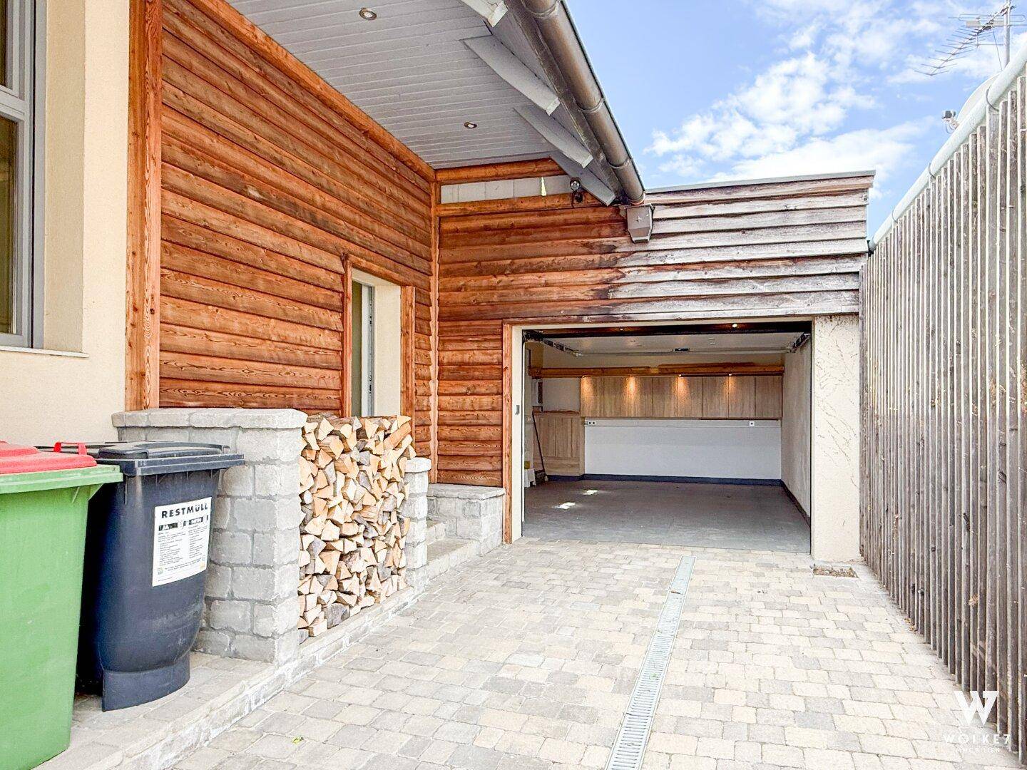 Garage mit Holzfassade, gepflasterter Einfahrt und gestapeltem Brennholz.