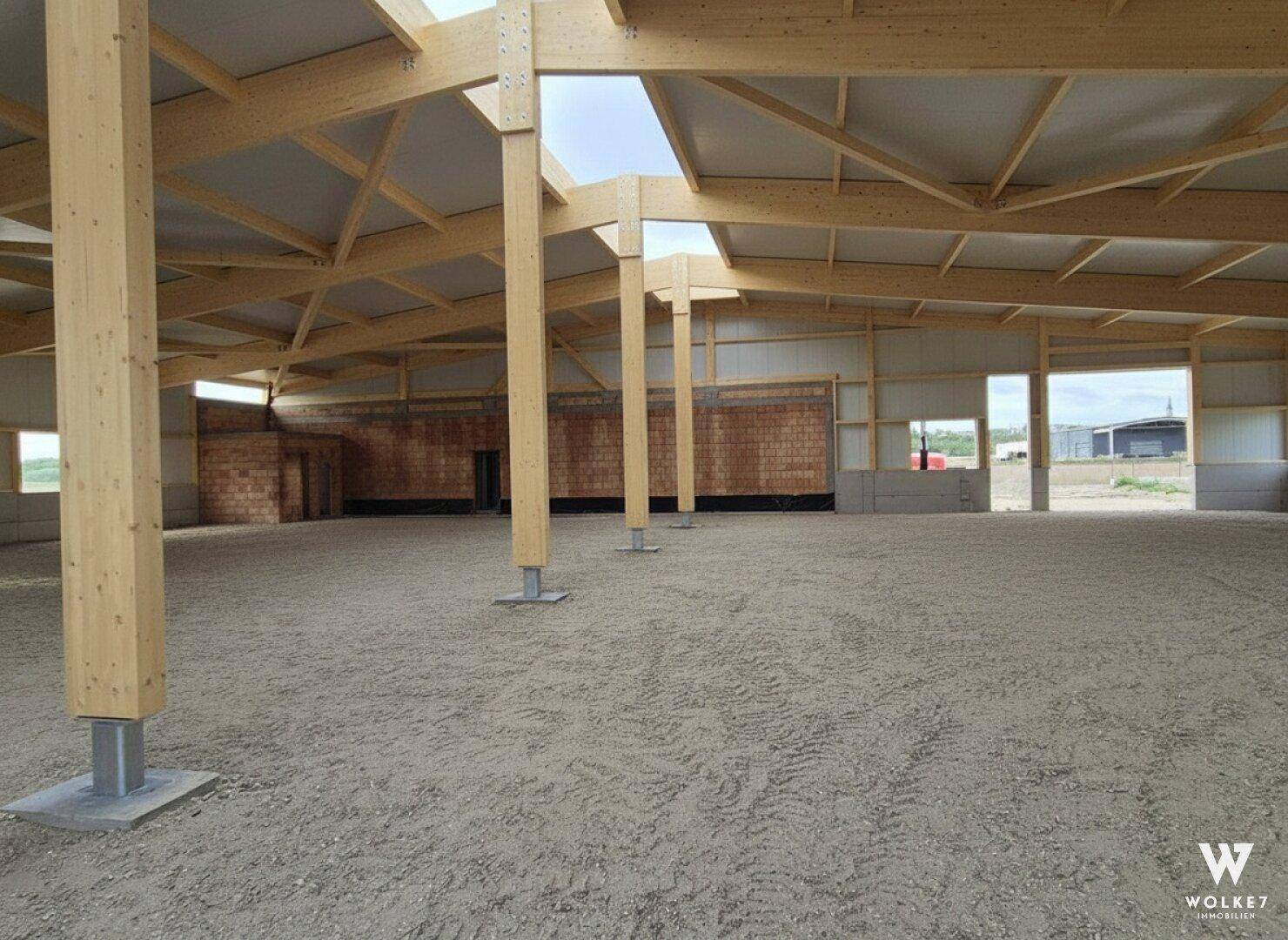 Innenansicht einer großen Halle mit Holzstützen und -balken sowie einem Sandboden während der Bauphase.