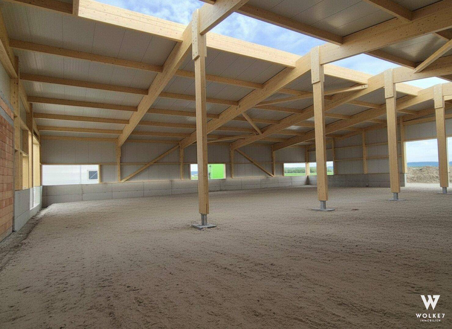 Geräumige Halle im Bau mit sichtbarer Holztragkonstruktion und großen Fensteröffnungen, die Licht hereinlassen.
