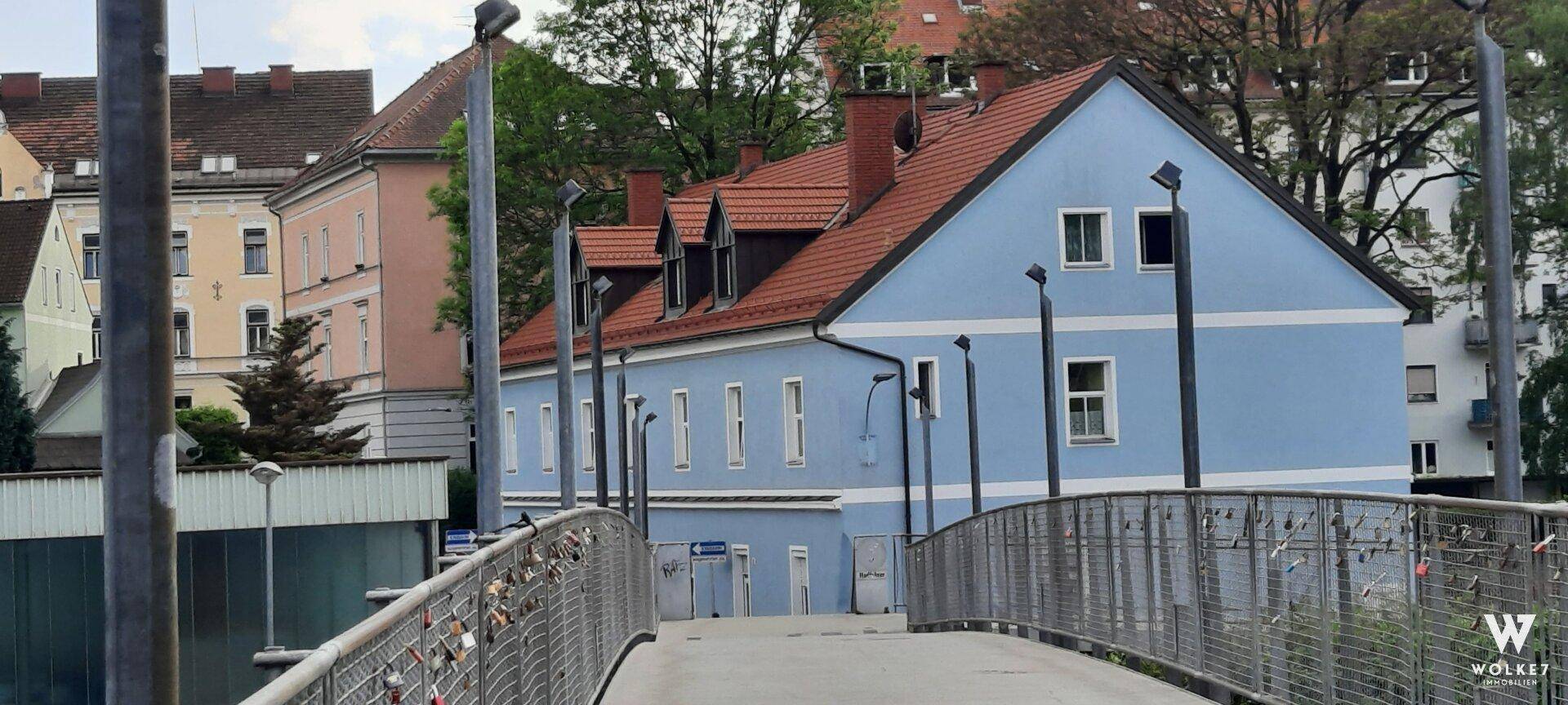 Blick auf das blaue Gebäude mit Ziegeldach von einer Fußgängerbrücke, umgeben von städtischer Bebauung.