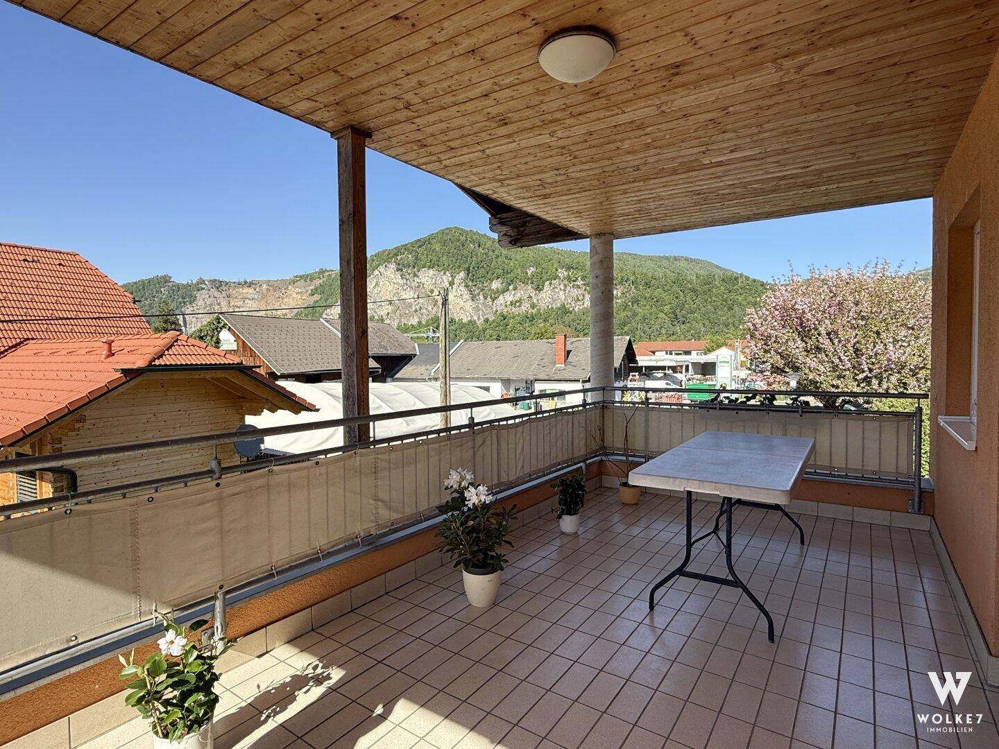Geräumiger überdachter Balkon mit Fliesenboden, Tisch und weitem Blick auf die umliegende Berglandschaft.