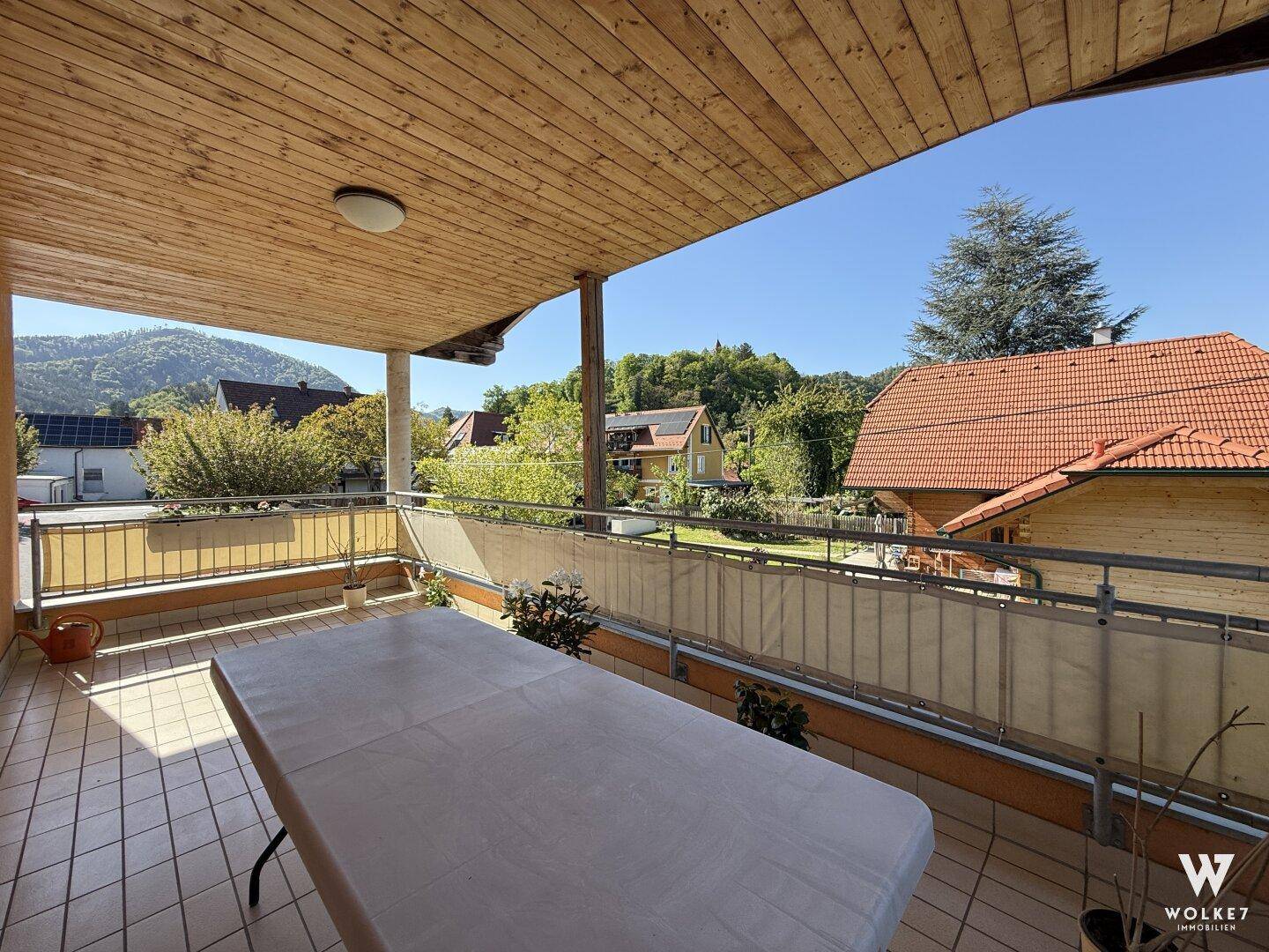 Heller überdachter Balkon mit Fliesenboden und weitem Blick auf die grüne Umgebung und Häuser.