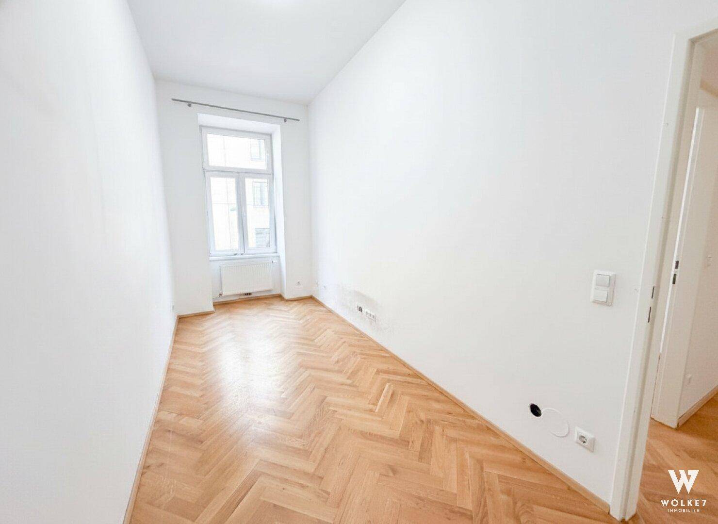 Helles, leeres Zimmer mit Fischgrätparkett und Fenster, ideal als Schlaf- oder Arbeitszimmer.