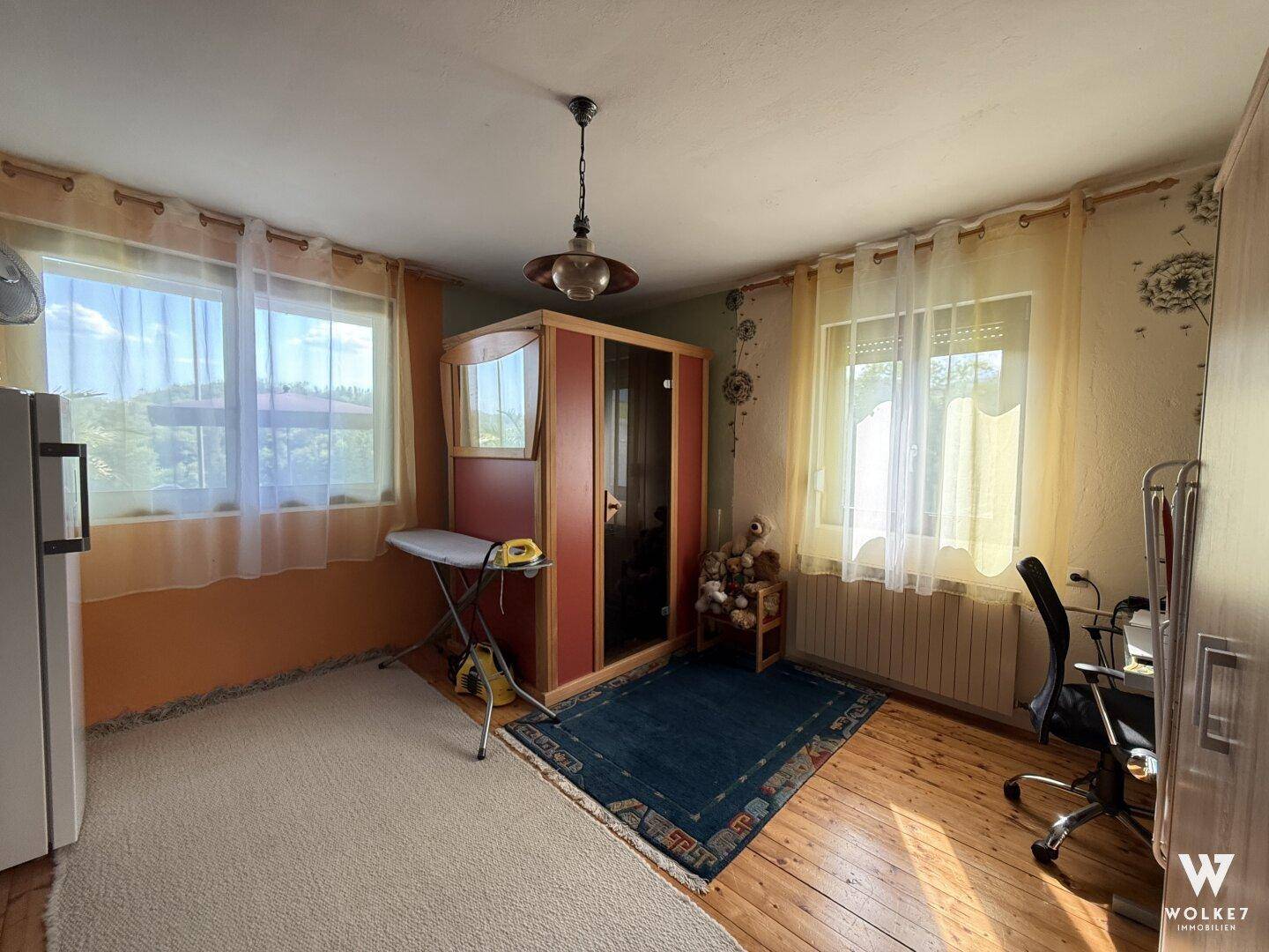 Helles Zimmer mit Holzboden, großem Fenster und einem Kleiderschrank mit Spiegel.