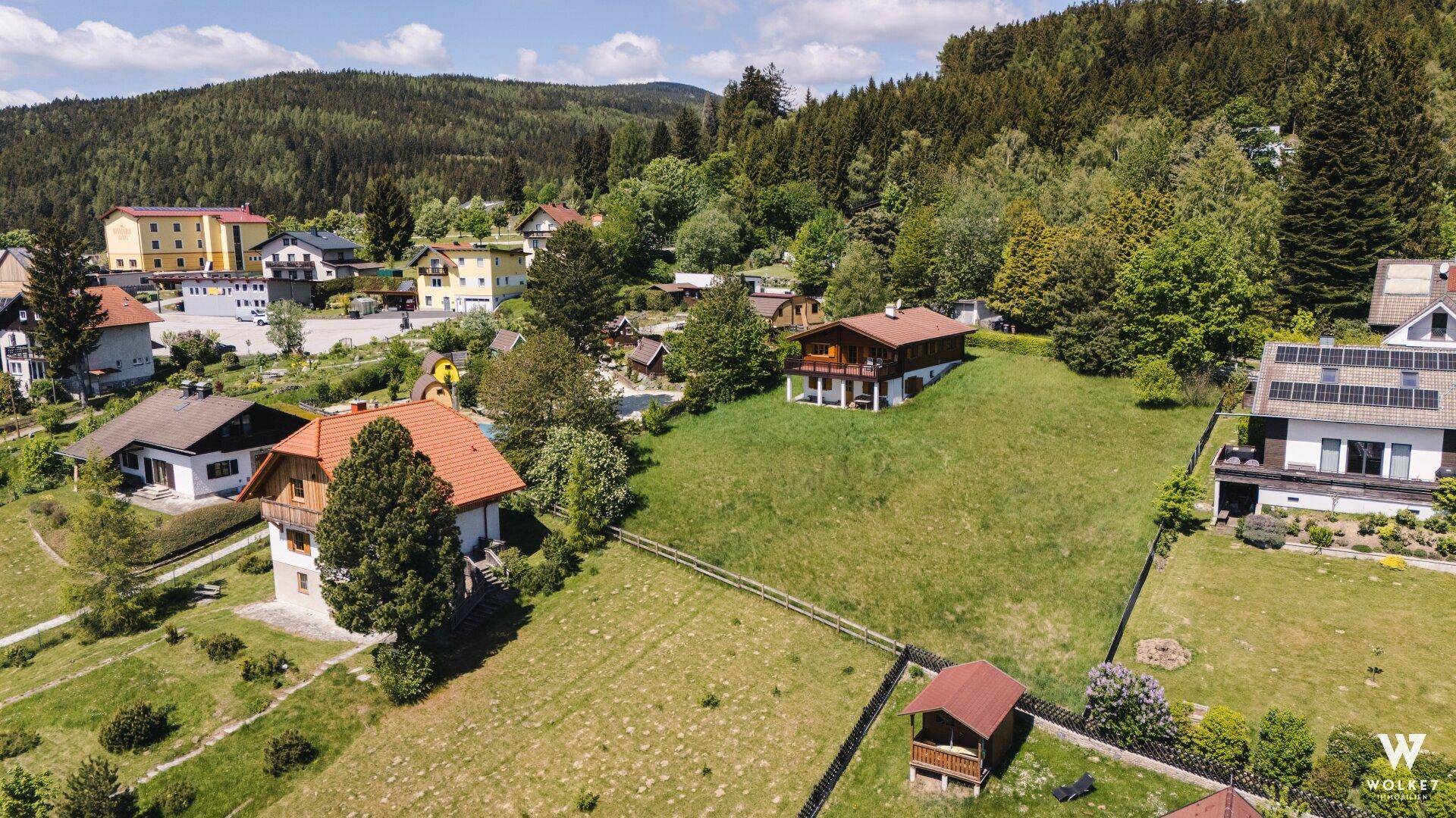 Luftaufnahme eines Wohngebiets mit mehreren Häusern, viel Grün und bewaldeten Hügeln im Hintergrund.