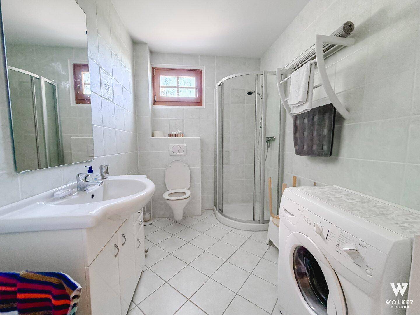 Badezimmer mit Duschkabine, Waschbecken, WC und Waschmaschine, hell gefliest.