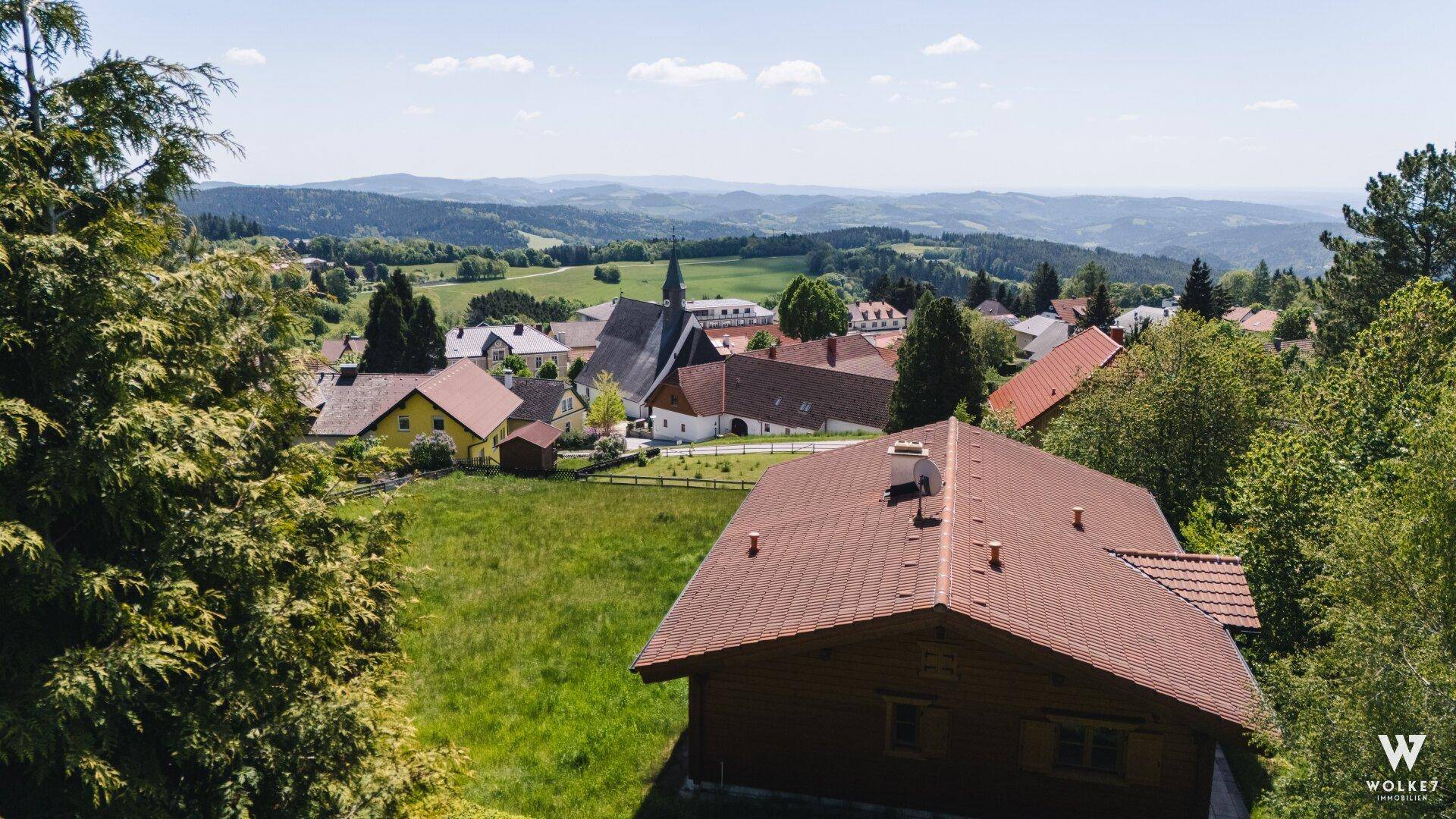 Panoramablick über ein malerisches Dorf mit Kirche und weiten grünen Hügeln unter blauem Himmel.