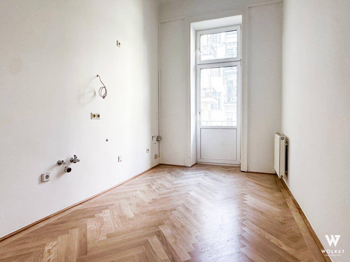Heller Raum mit Fischgrätparkett, Balkontür und sichtbaren Installationen für zukünftige Küchen- oder Badnutzung.