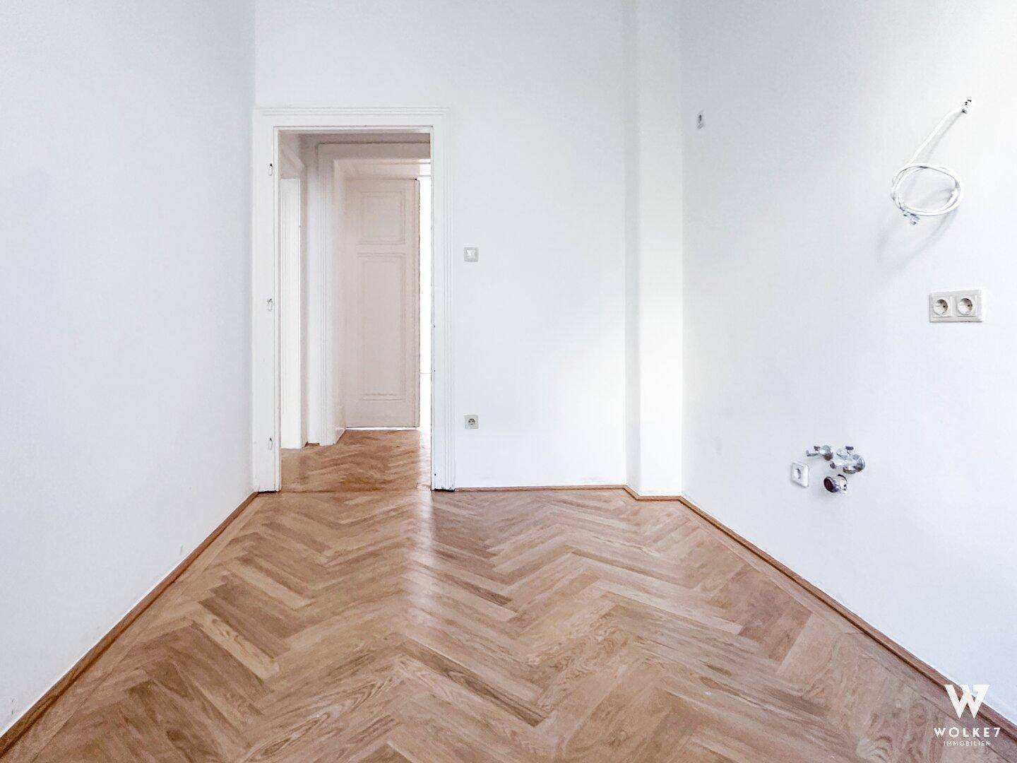 Leerer Raum mit Fischgrätparkett, weißen Wänden und einem offenen Durchgang zu einem angrenzenden Zimmer.