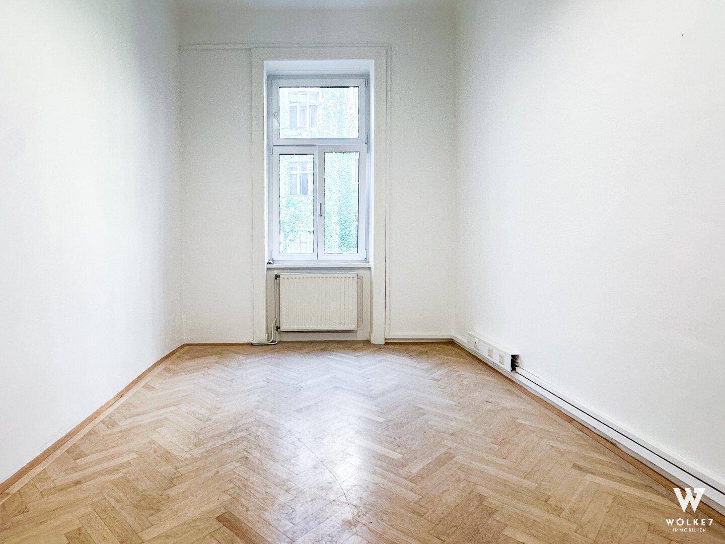 Heller, leerer Raum mit weißen Wänden, klassischem Fischgrätparkett und einem Fenster, das Tageslicht hereinlässt.