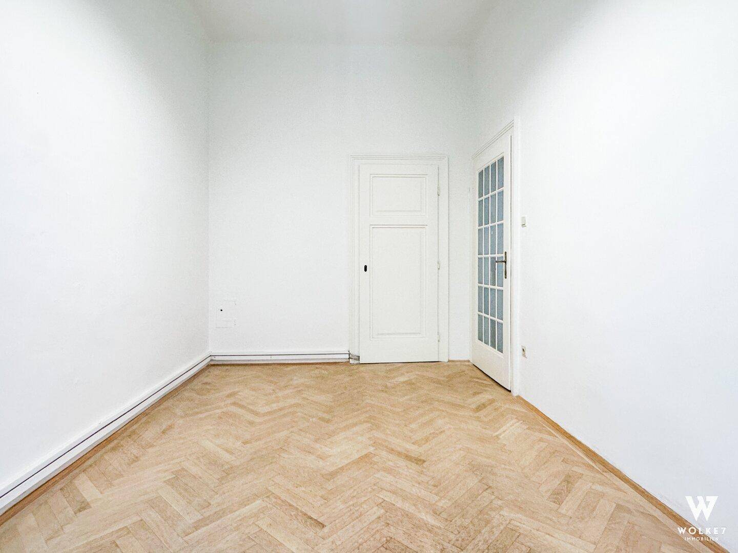 Leerer Raum mit elegantem Fischgrätparkett und zwei weißen Türen, eine davon mit dekorativen Glaseinsätzen.