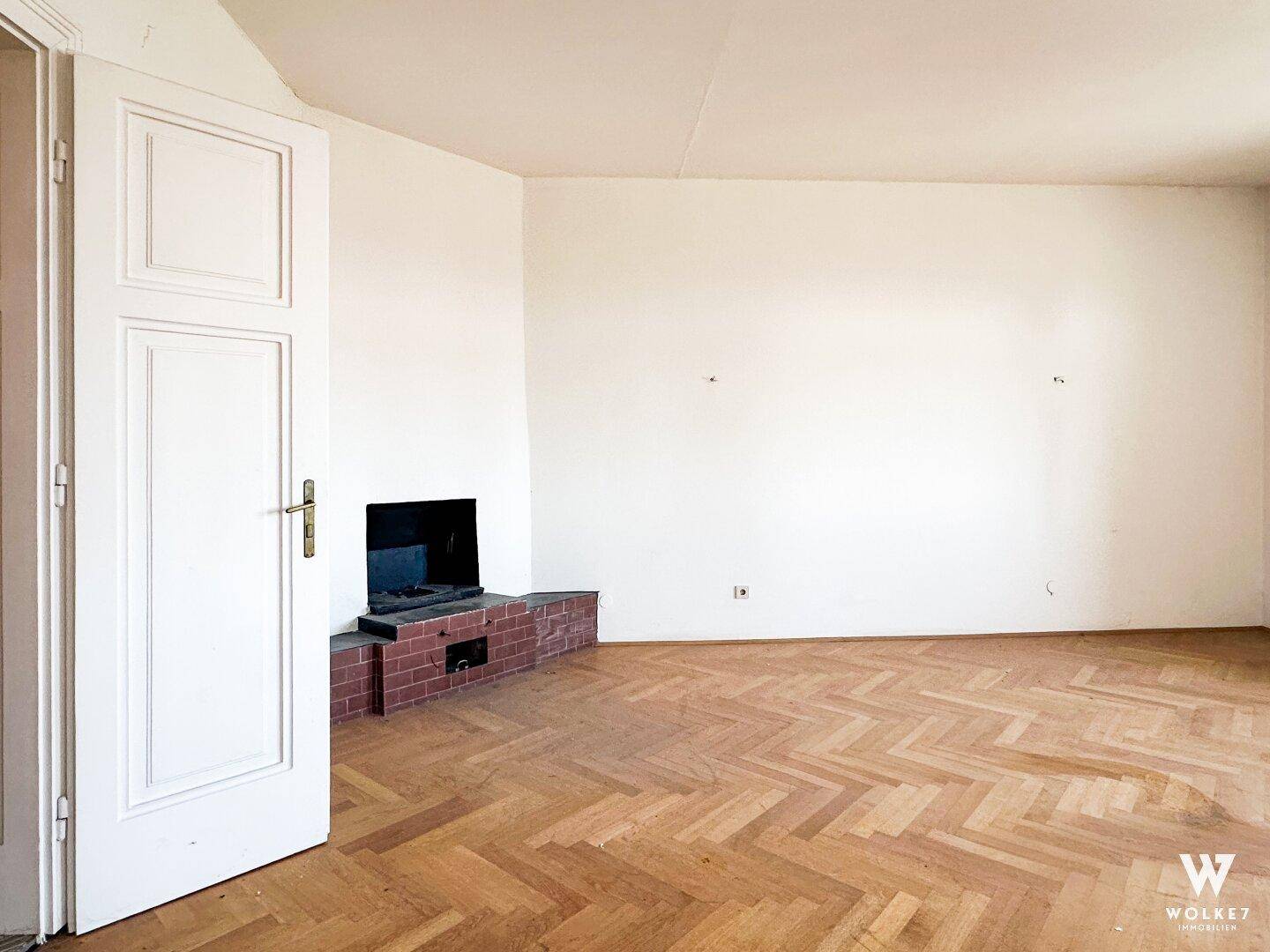 Helles Zimmer mit Fischgrätparkett und einem gemauerten Kamin in der Ecke, einladend und geräumig.