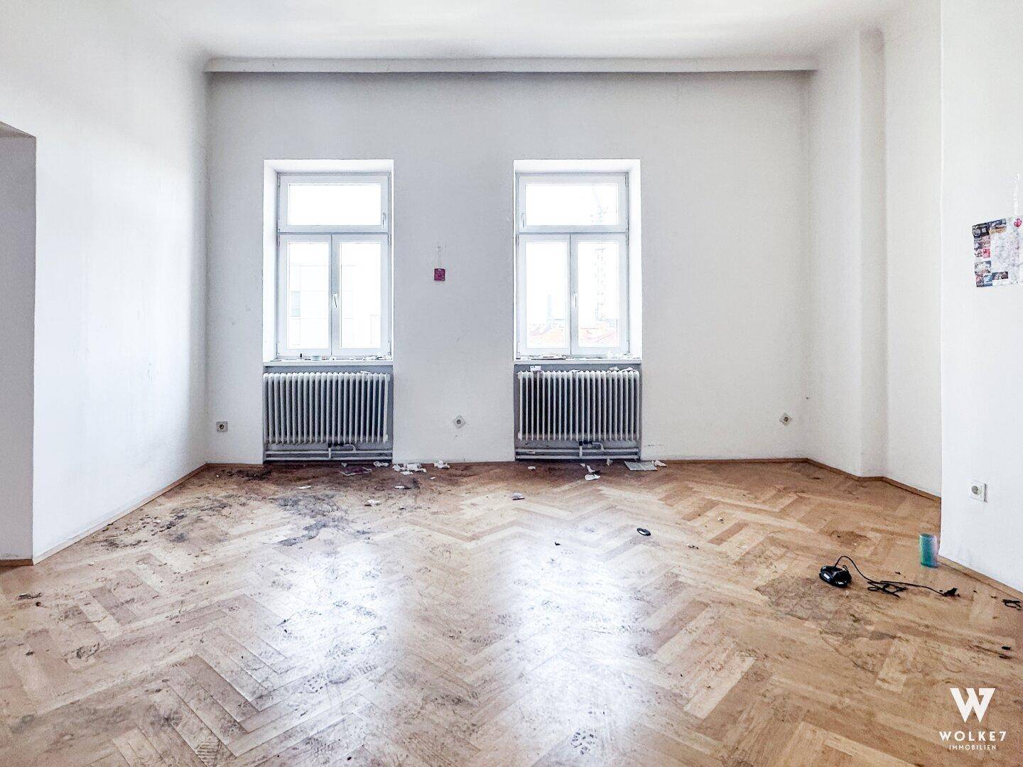 Renovierungsbedürftiges Zimmer mit Fischgrätparkett und zwei Fenstern, bereit für eine Neugestaltung.