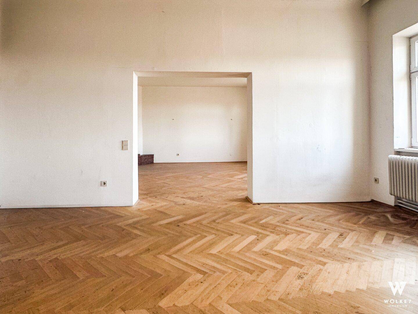 Geräumiges Zimmer mit Fischgrätparkett und offenem Durchgang zu einem weiteren Raum.