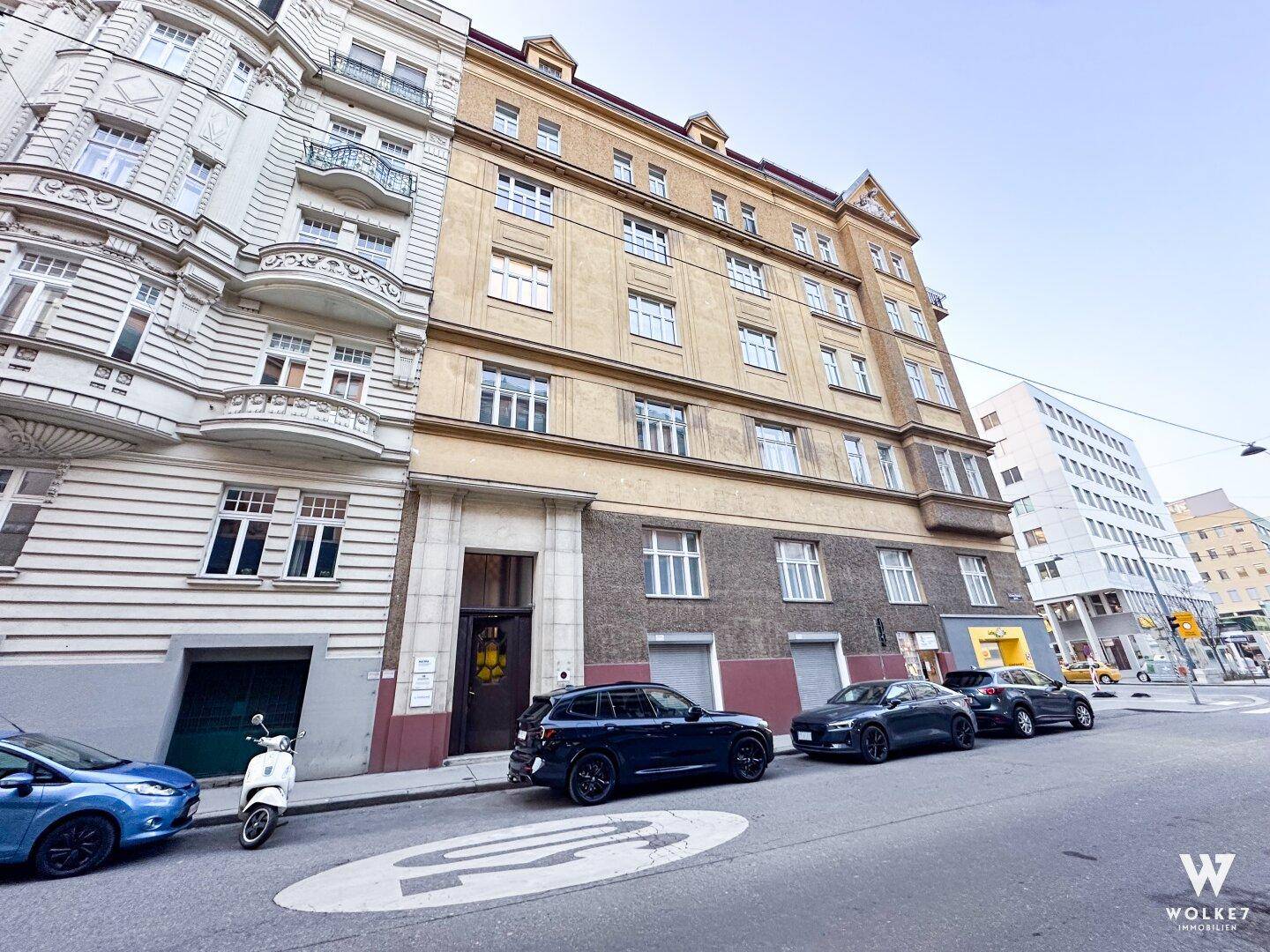 Historisches Gebäude mit klassischer Fassade und parkenden Autos auf der Straße davor.