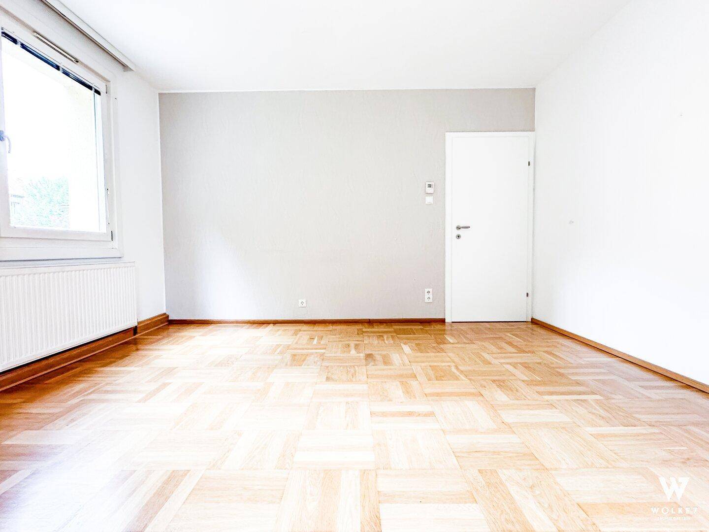 Heller, leerer Raum mit Parkettboden und einem Fenster, das viel Tageslicht hereinlässt.