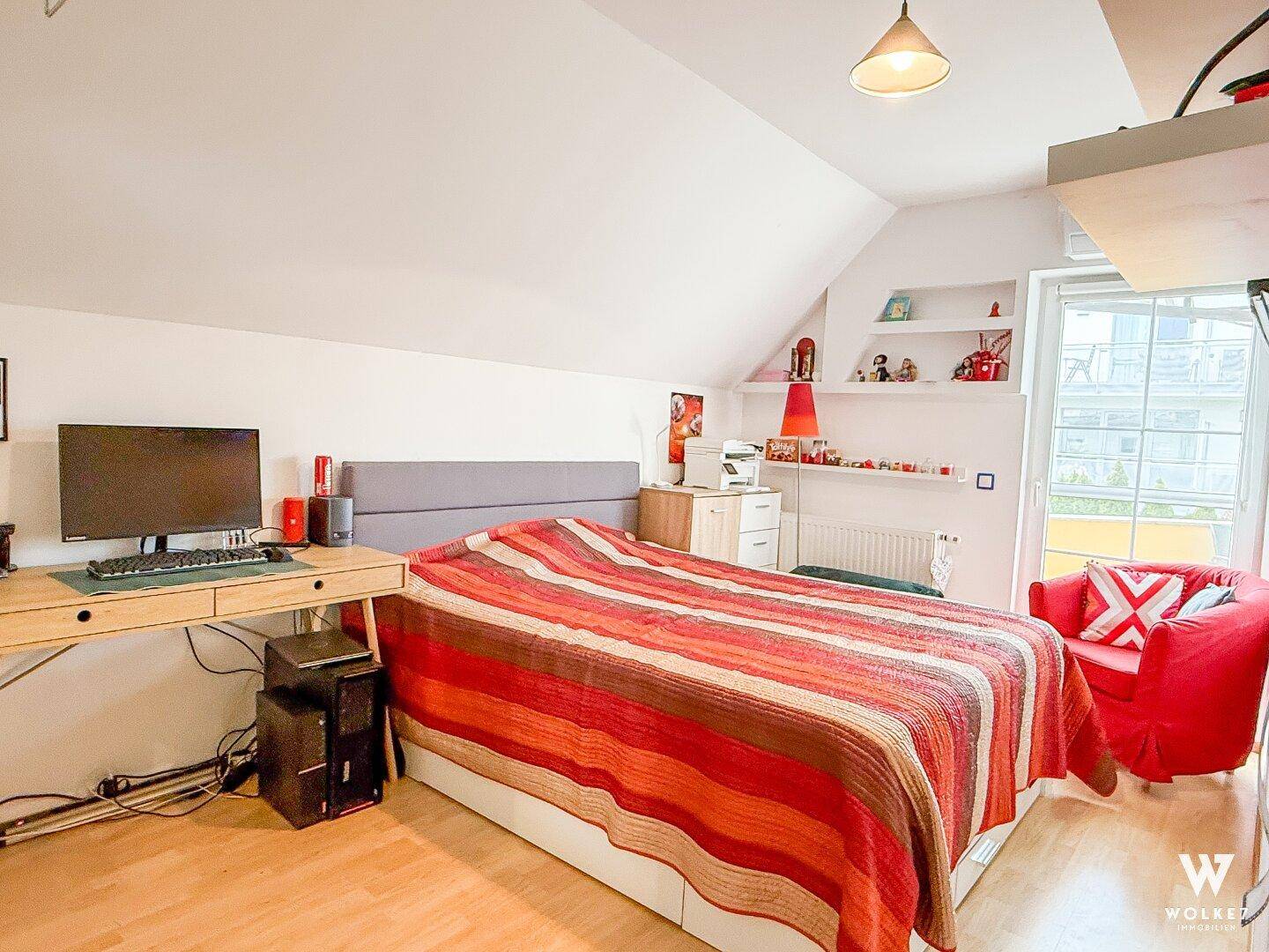 Helles Schlafzimmer mit Schreibtisch, Bett und farbenfroher Bettwäsche unter der Dachschräge.