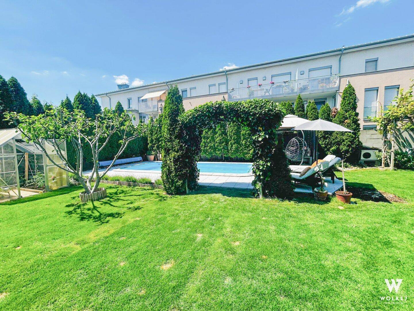 Weitläufiger Garten mit Pool, Liegestühlen und einem Gewächshaus unter blauem Himmel.