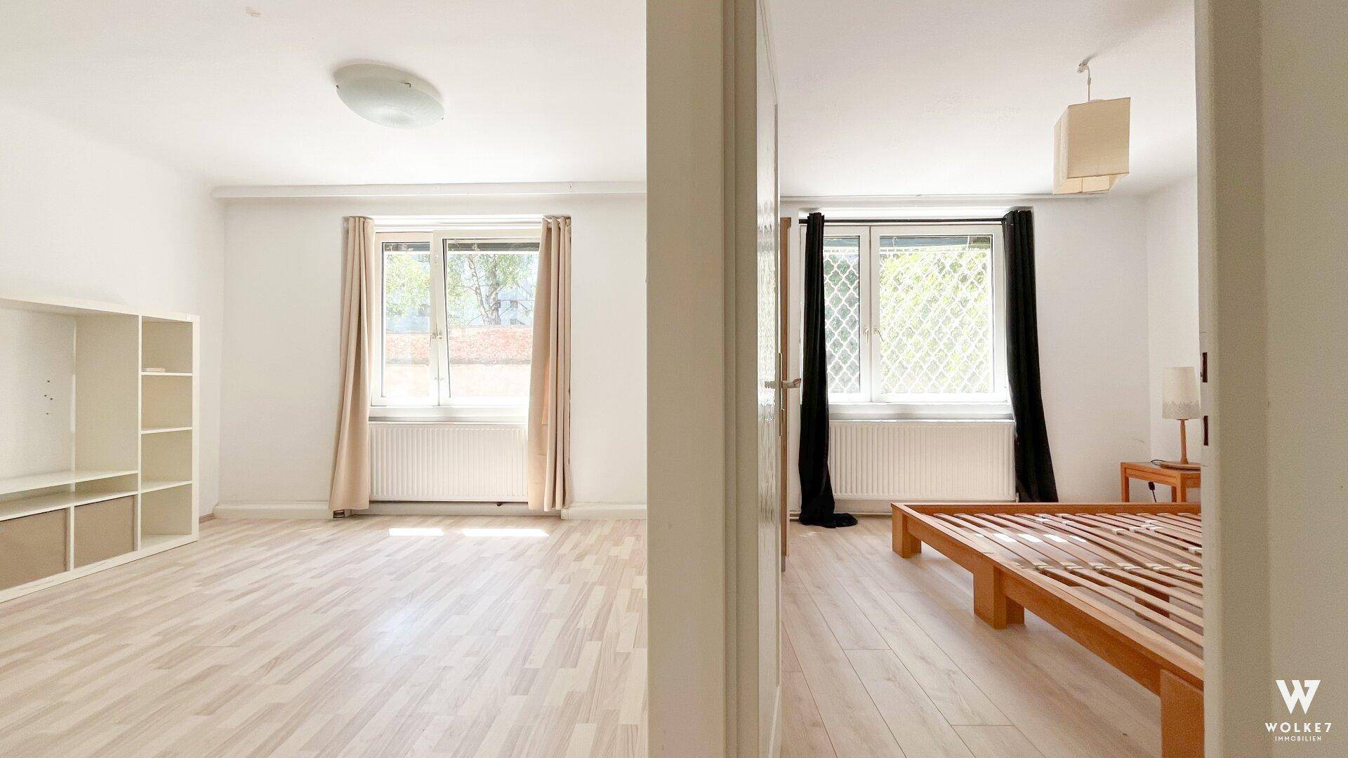 Blick von einem leeren Raum in ein Schlafzimmer mit Bettrahmen und Fenster, beide mit Laminatboden.