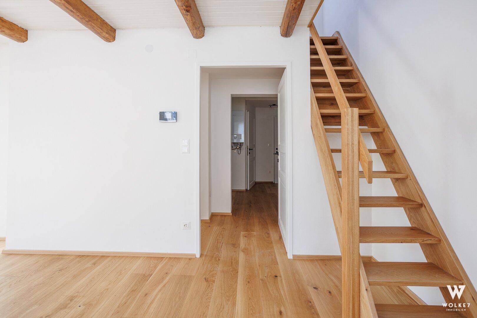 Heller Flurbereich mit Holztreppe, Holzboden und sichtbaren Deckenbalken, führt zu weiteren Räumen.