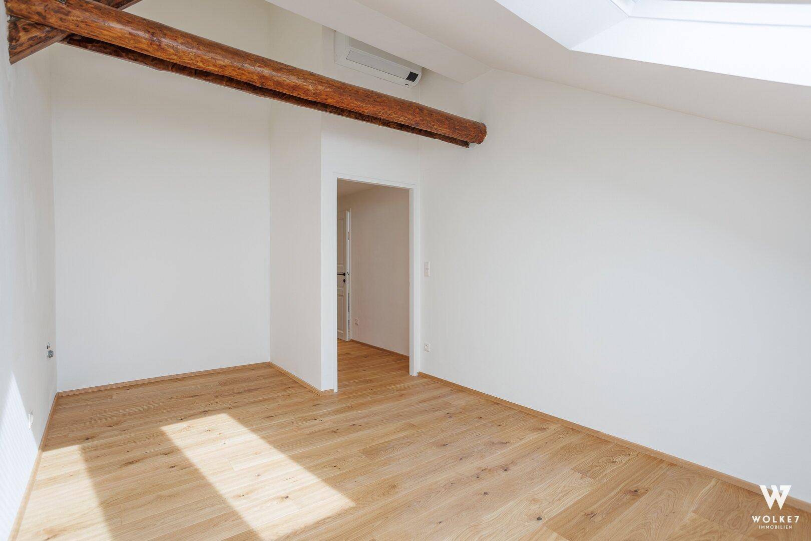 Leeres Zimmer im Dachgeschoss mit Holzboden, sichtbarem Holzbalken und viel natürlichem Licht.