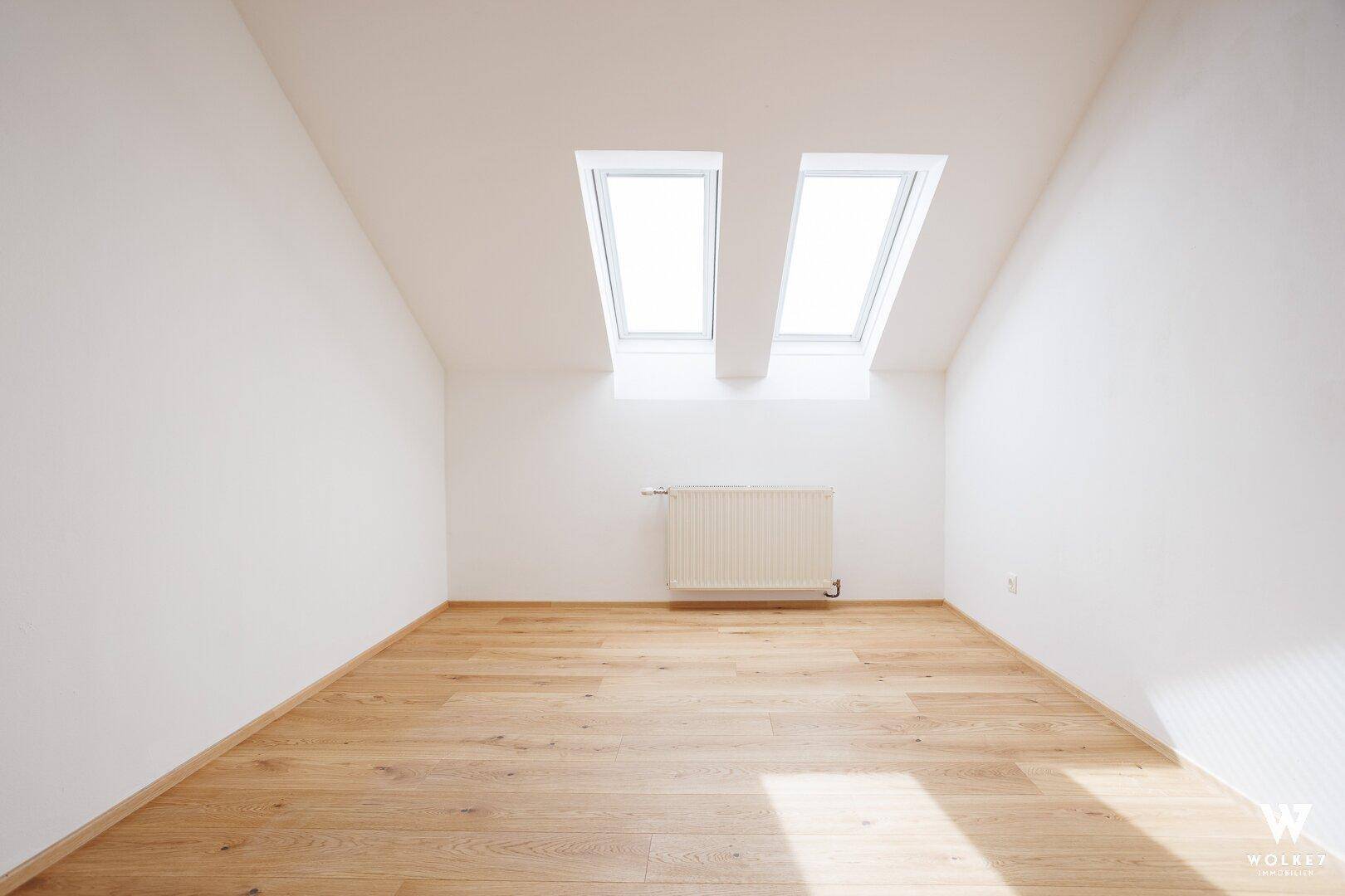 Helles Zimmer im Dachgeschoss mit Holzboden, Heizkörper und zwei großen Dachfenstern.