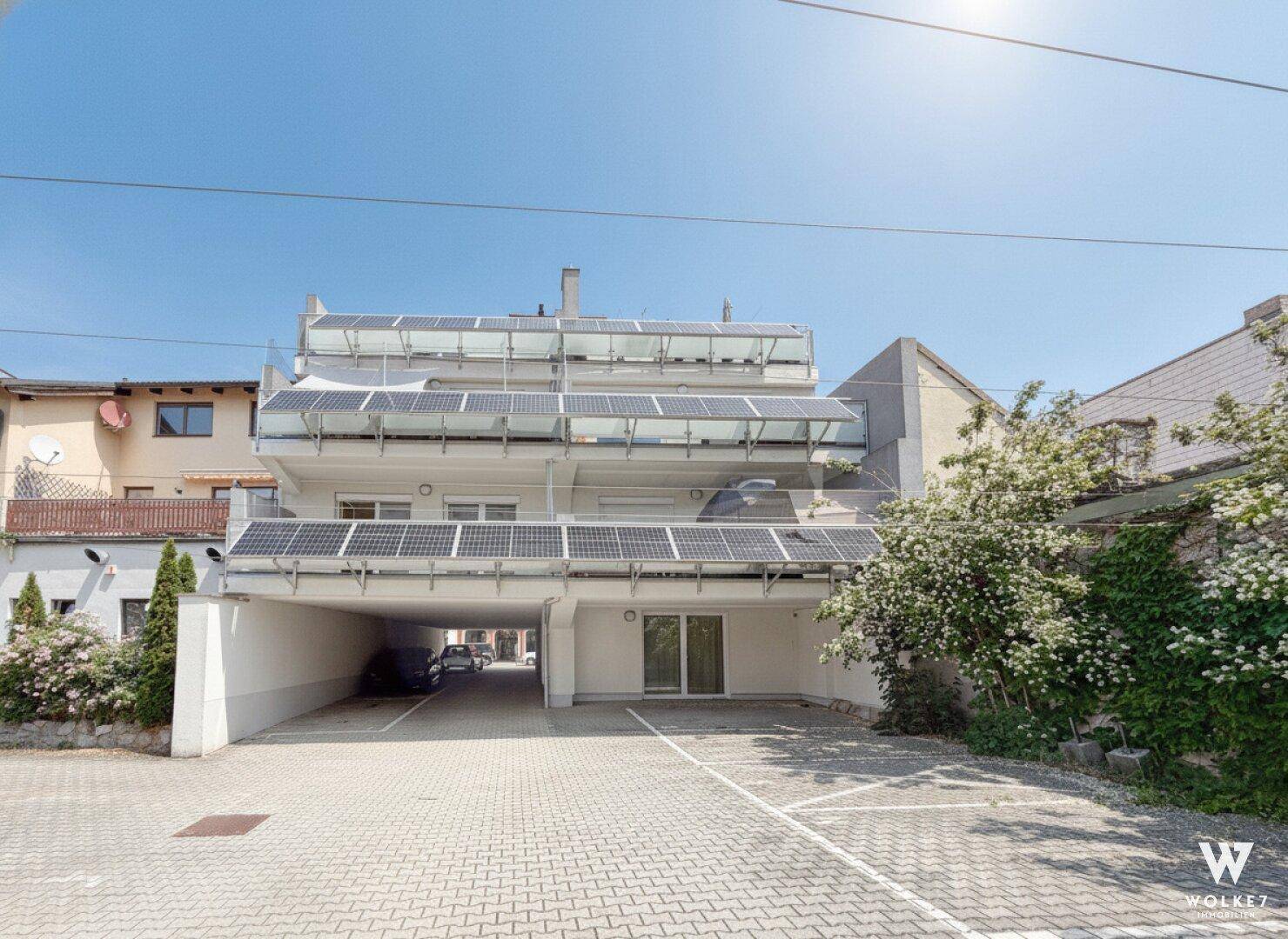 Sonnenbeschienene Rückseite des Gebäudes mit Balkonen, Solarmodulen und gepflastertem Parkplatz.