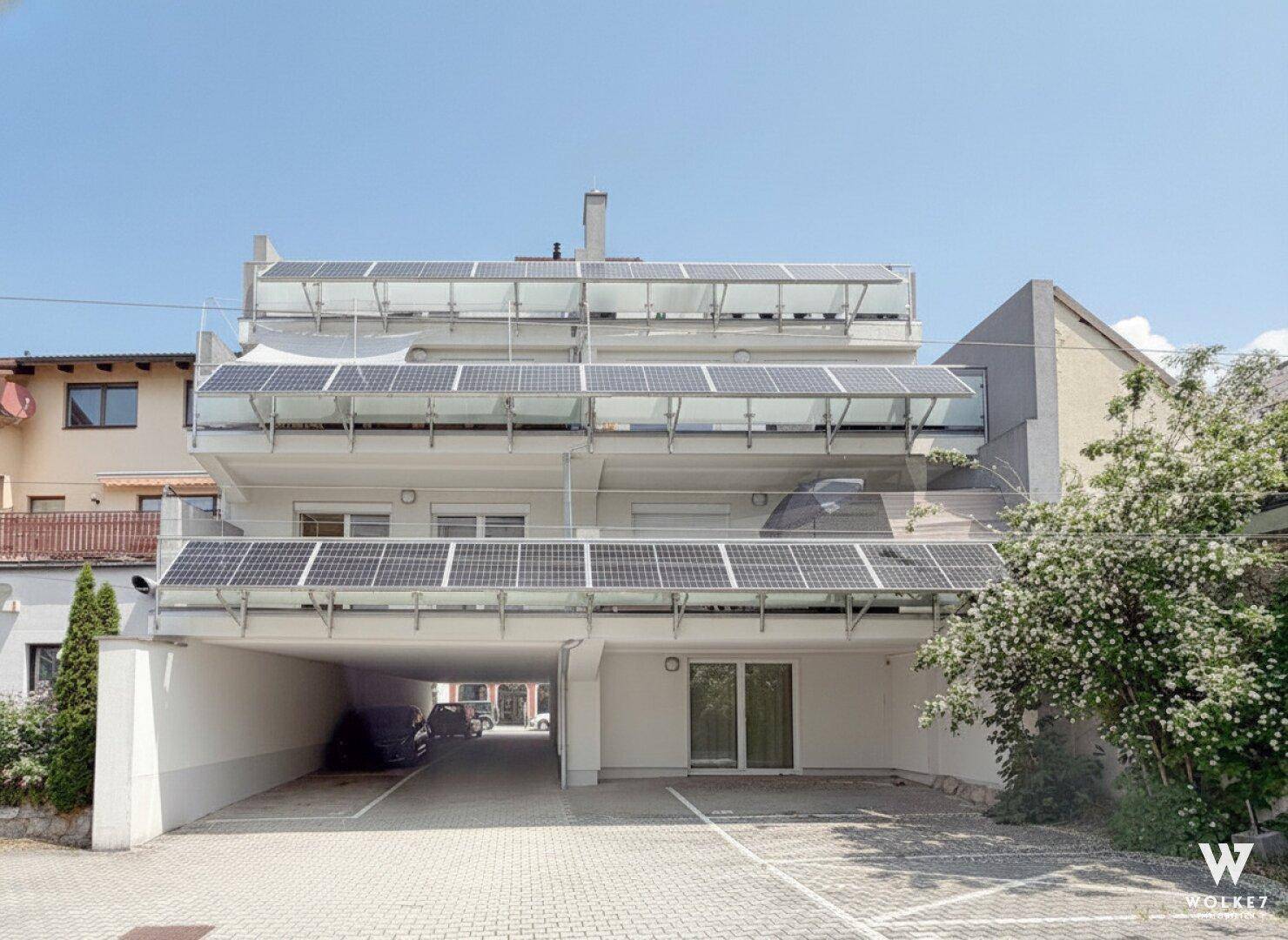 Gebäuderückseite mit mehreren Balkonen, Photovoltaik-Anlage und gepflasterten Stellplätzen.