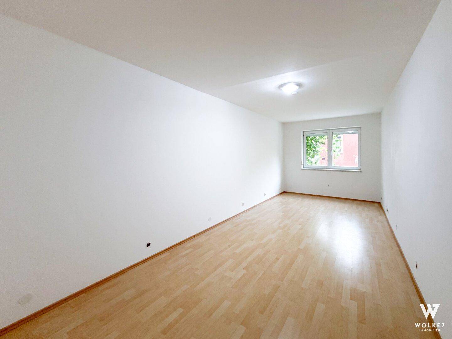 Geräumiger, leerer Raum mit hellem Laminatboden und einem Fenster mit Grünblick.