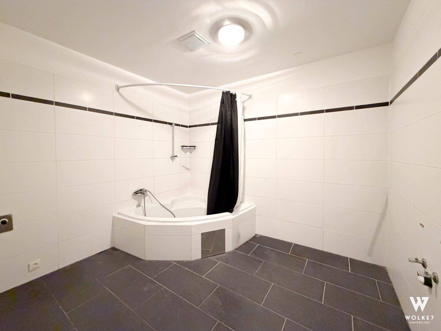 Helles Badezimmer mit weißen Fliesen, dunklem Boden und einer Eckbadewanne mit Duschvorhang.