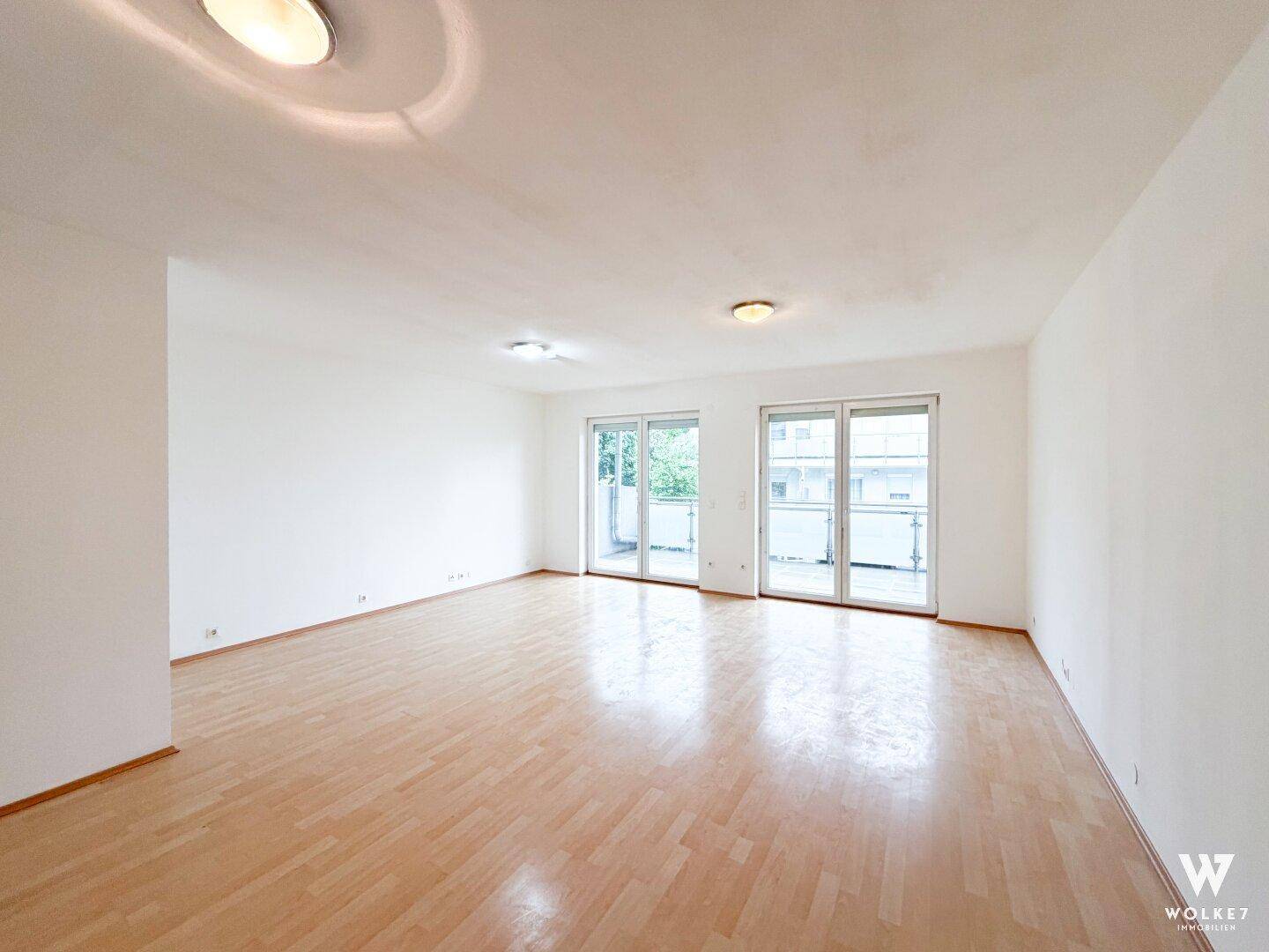 Heller Wohnraum mit Laminatboden und zwei großen Fenstertüren zum Balkon.