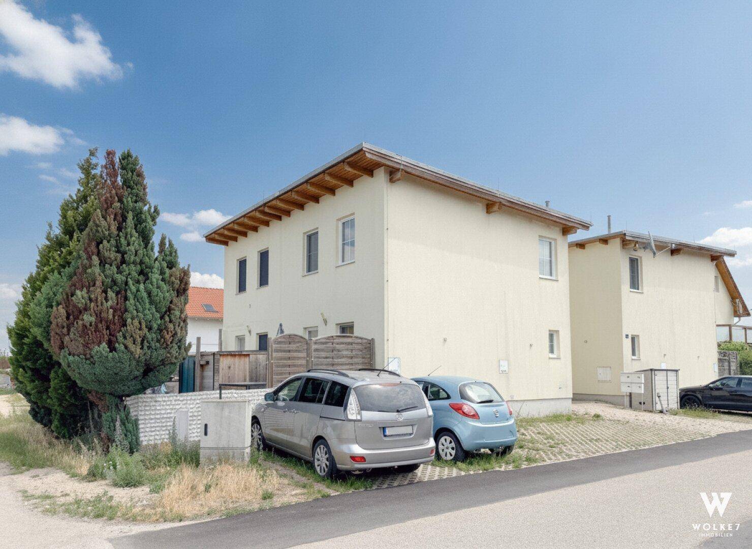 Ansicht eines Mehrfamilienhauses von der Seite mit Parkplätzen und einem hohen Nadelbaum im Vordergrund.