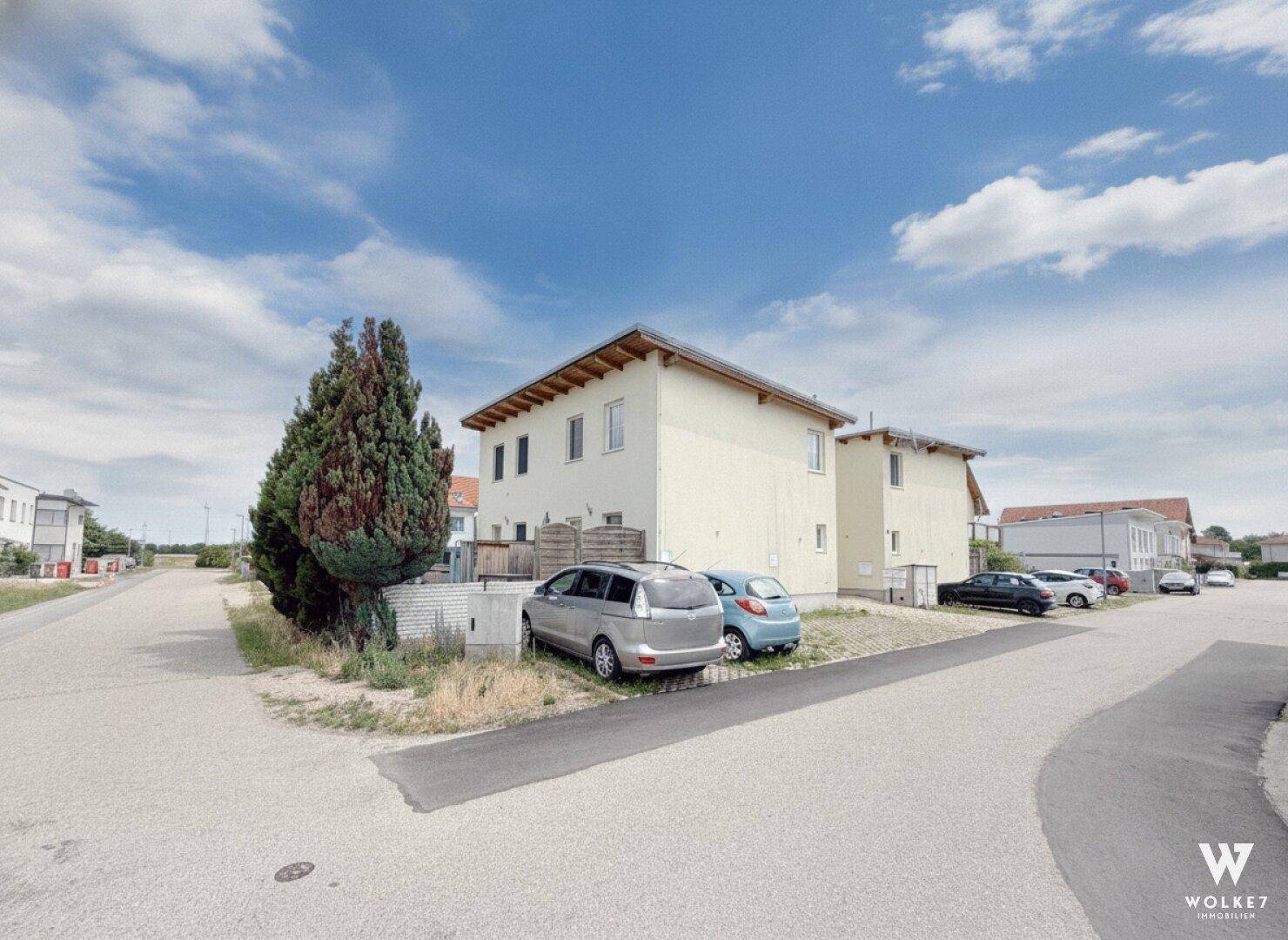 Weitläufige Ansicht eines Mehrfamilienhauses mit Parkplätzen und einem großen Baum unter blauem Himmel.