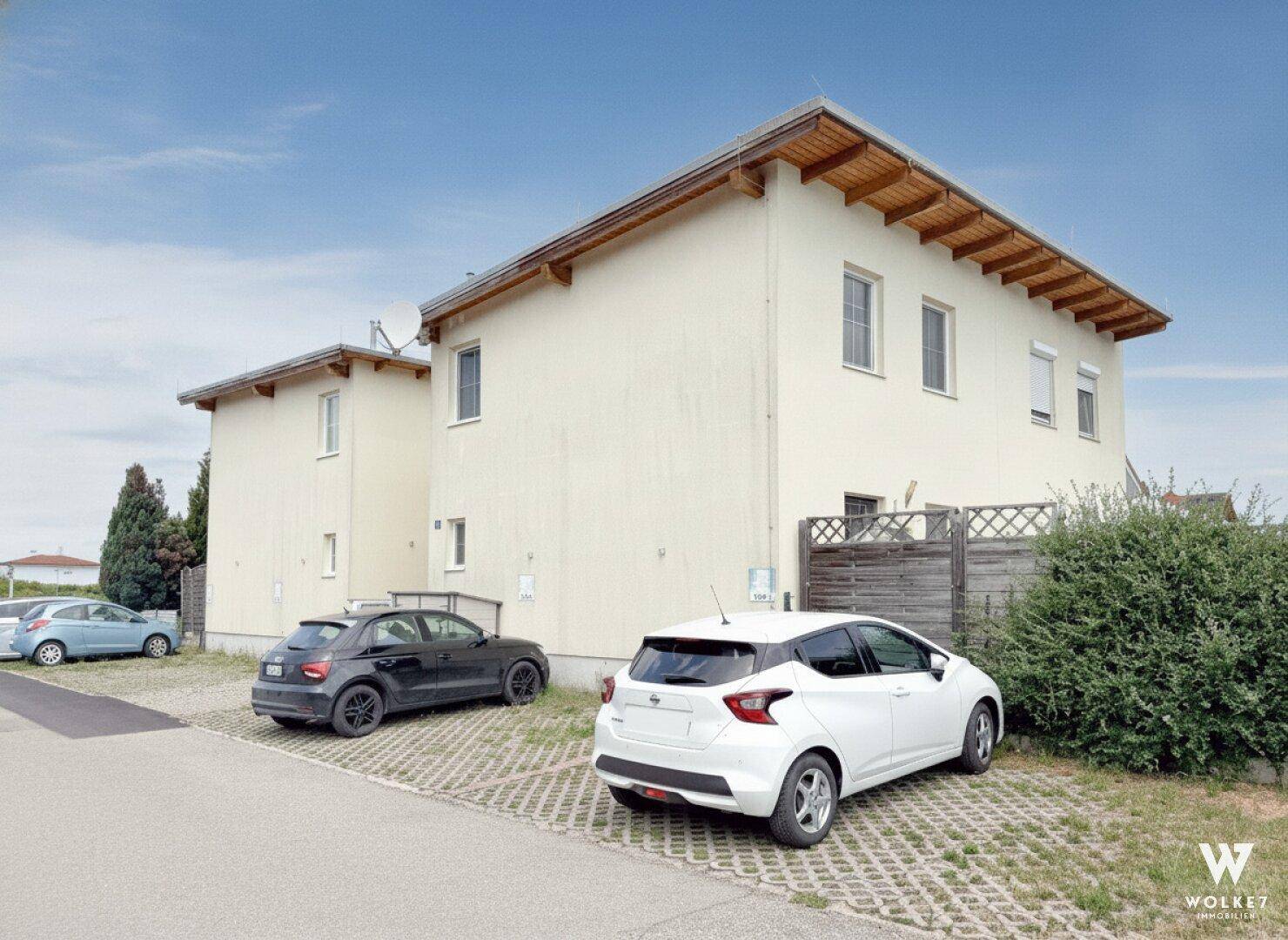 Seitenansicht eines Mehrfamilienhauses mit heller Fassade, Holzsparren und gepflasterten Parkflächen.