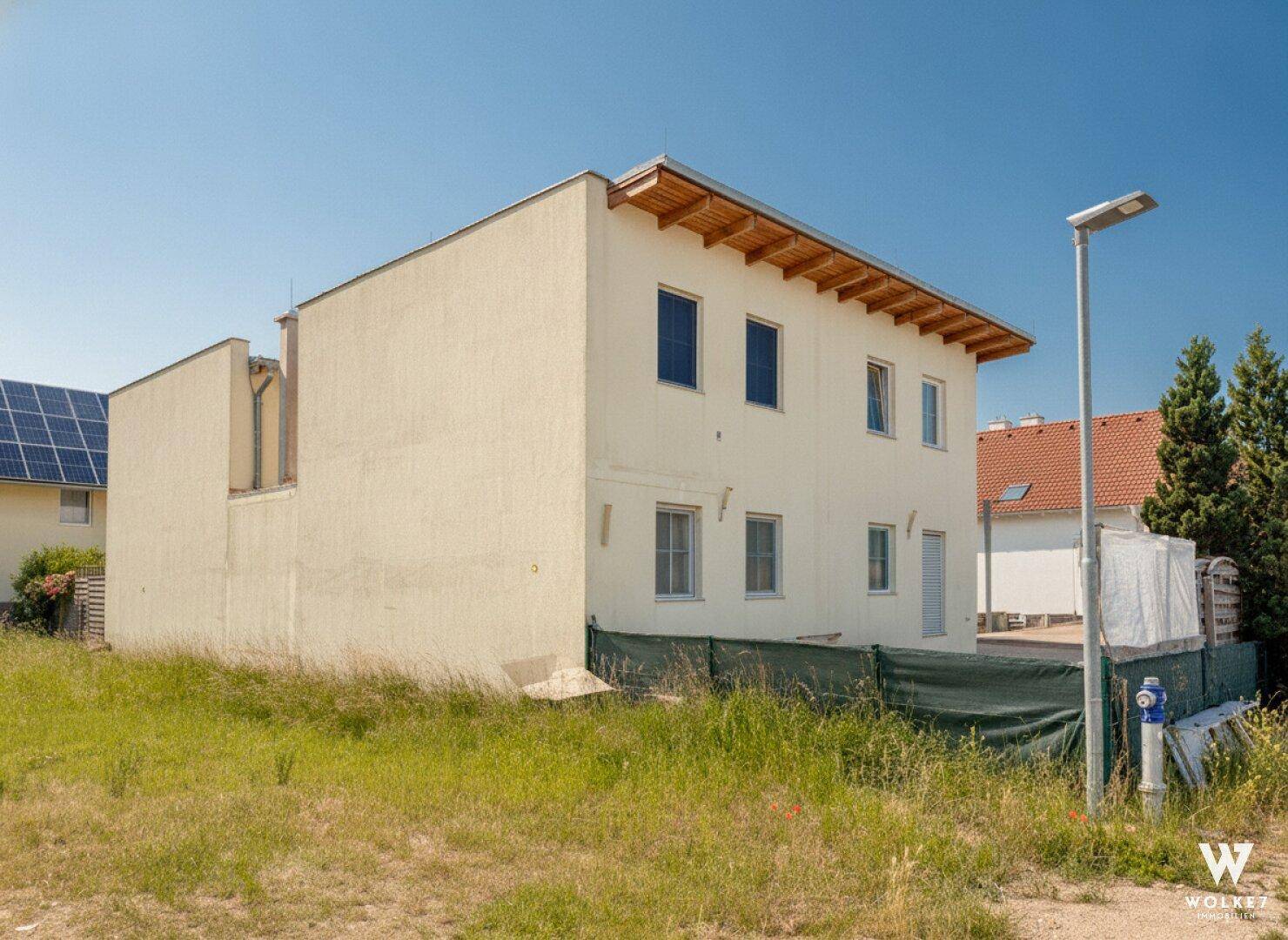 Seitenansicht eines Mehrfamilienhauses mit heller Fassade und angrenzendem unbebautem Grünland.
