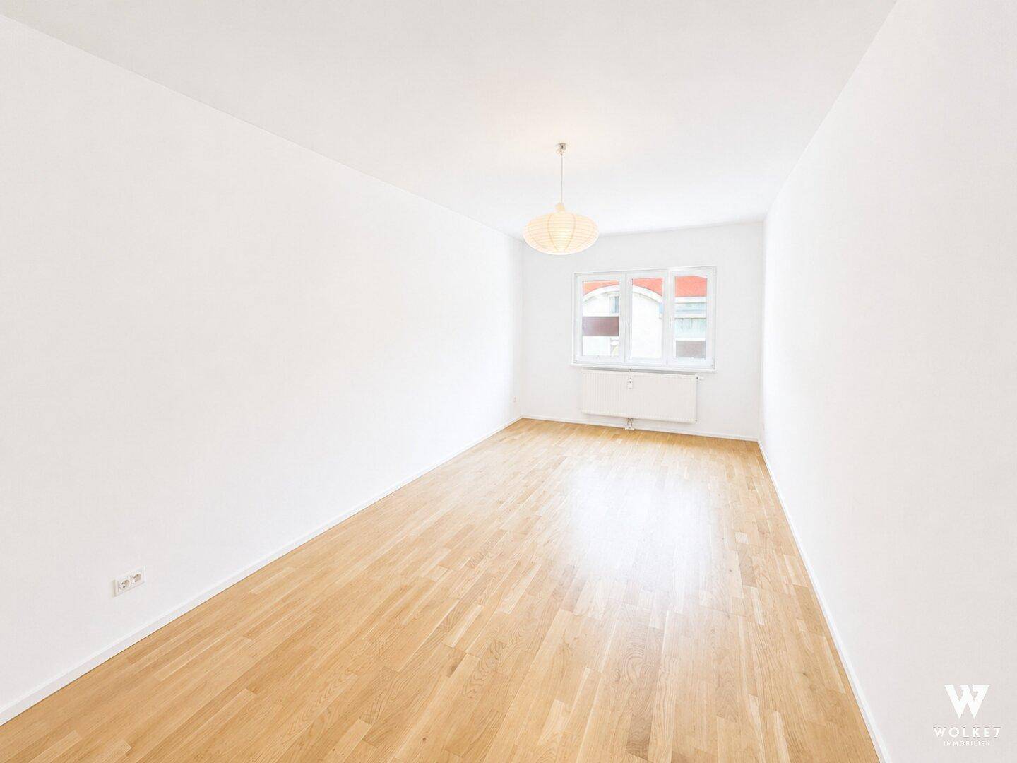 Geräumiges, leeres Zimmer mit hellem Parkettboden und einem großen Fenster, das viel Licht hereinlässt.