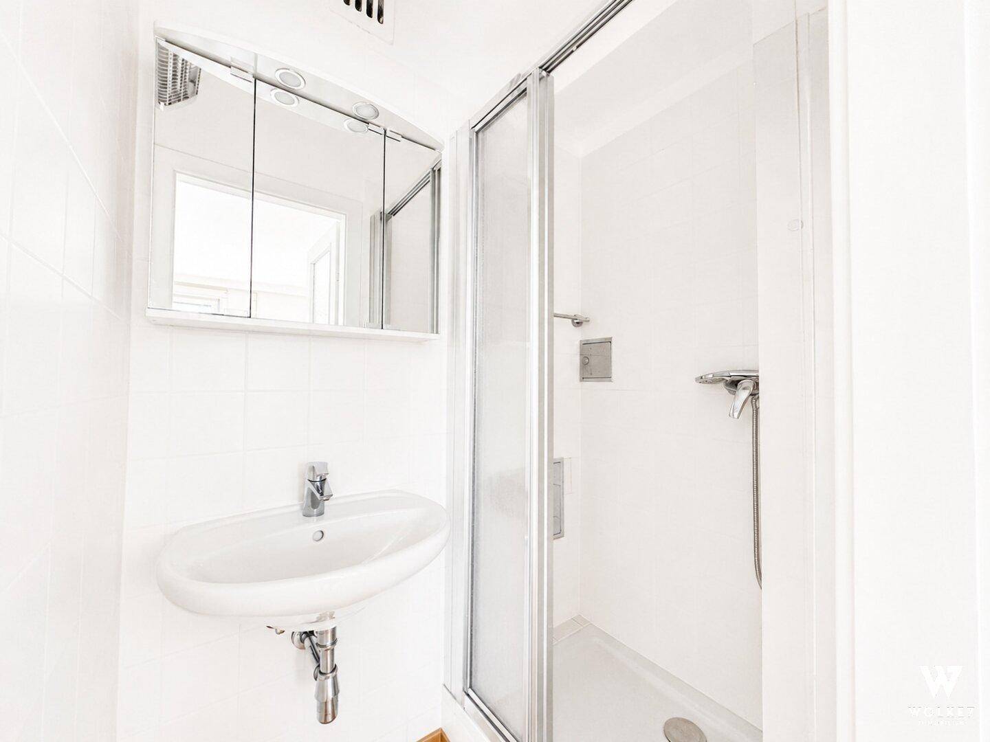 Kleines Badezimmer mit Dusche, Waschbecken und Spiegelschrank, hell gefliest.