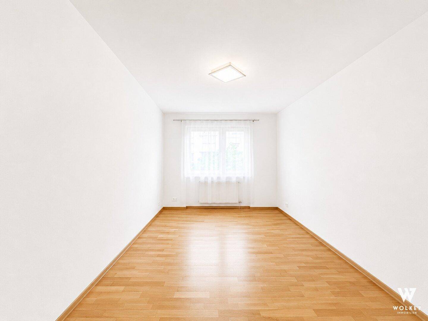 Geräumiges, leeres Schlafzimmer mit hellem Parkettboden und einem großen Fenster, das den Raum erhellt.