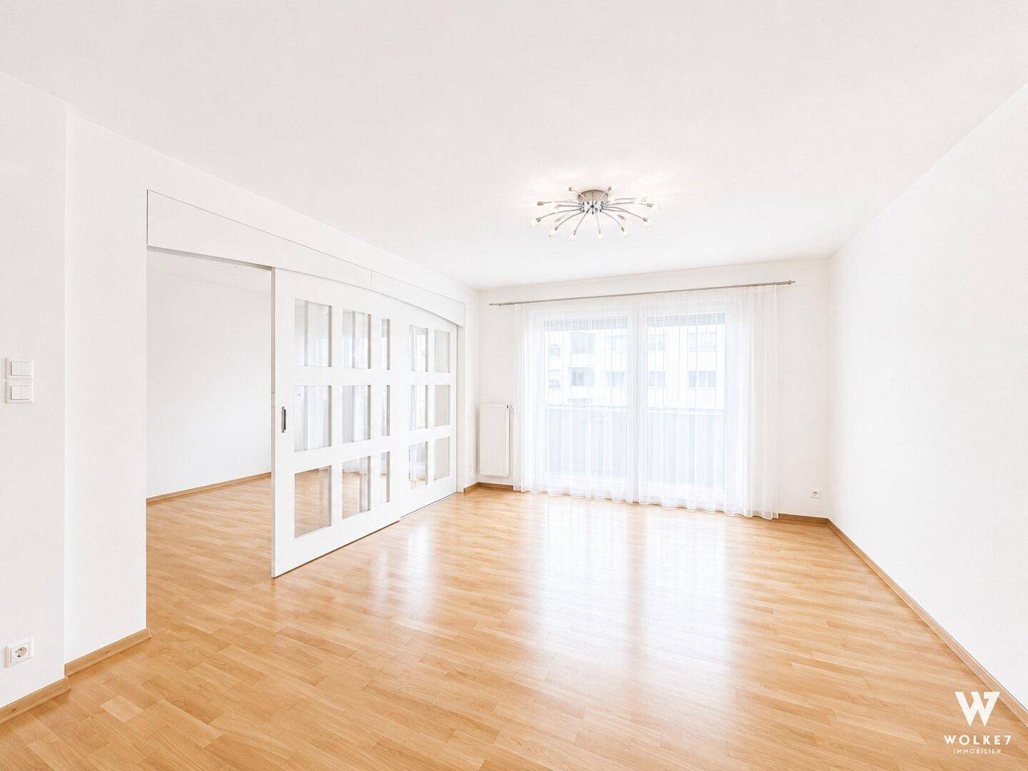 Geräumiges, leeres Wohnzimmer mit hellem Parkettboden und großen Fenstern, die viel Tageslicht hereinlassen.