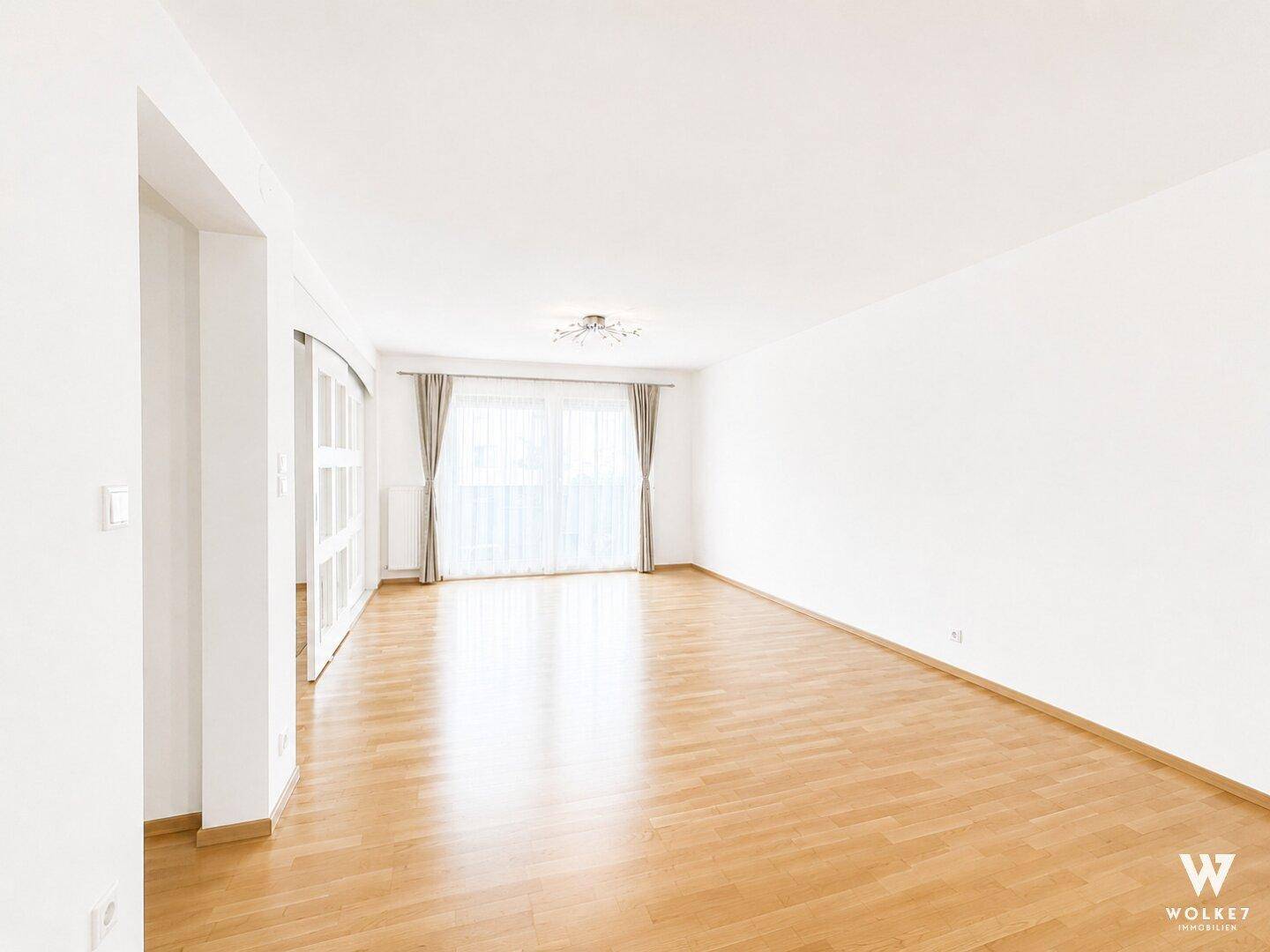 Weitläufiges, leeres Wohnzimmer mit Parkettboden und großen Fenstern, die den Raum hell erscheinen lassen.