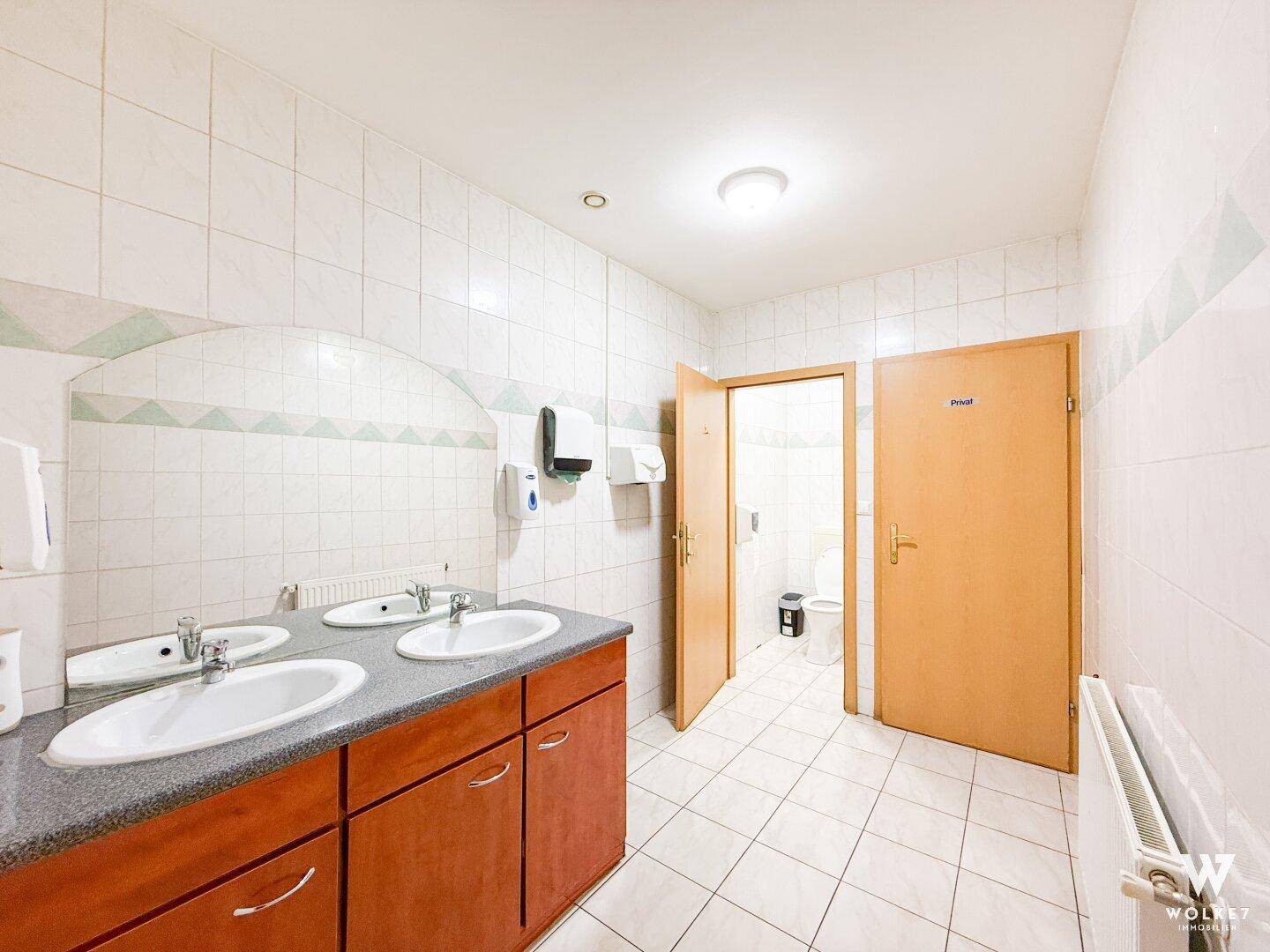 Sauberer öffentlicher Waschraum mit mehreren Waschbecken, großen Spiegeln und zwei Toilettenkabinen.