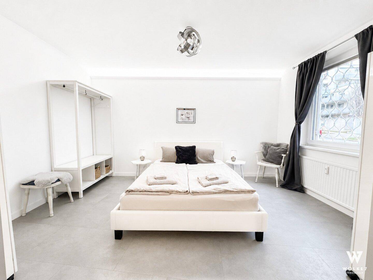 Schlichtes Schlafzimmer mit Doppelbett, offenem Kleiderschrank und großem Fenster.
