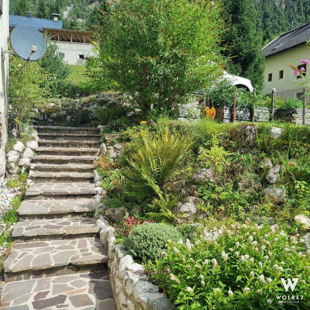 Steintreppe führt durch einen terrassierten Garten mit vielfältiger Bepflanzung und Bergblick im Hintergrund.