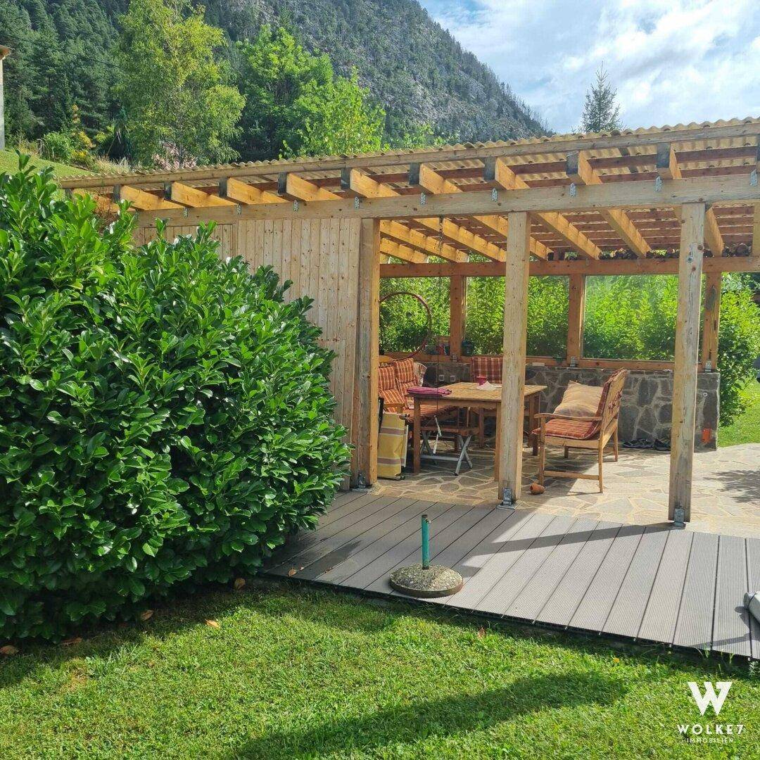 Gemütlicher Essbereich im Freien unter einer Holzpergola, umgeben von Rasen und mit Bergblick.