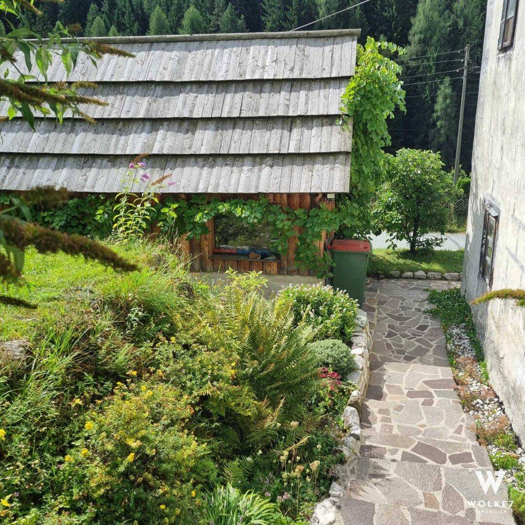 Steinweg und Treppe führen an einem kleinen Holzschuppen mit Schindeldach und üppiger Vegetation vorbei.